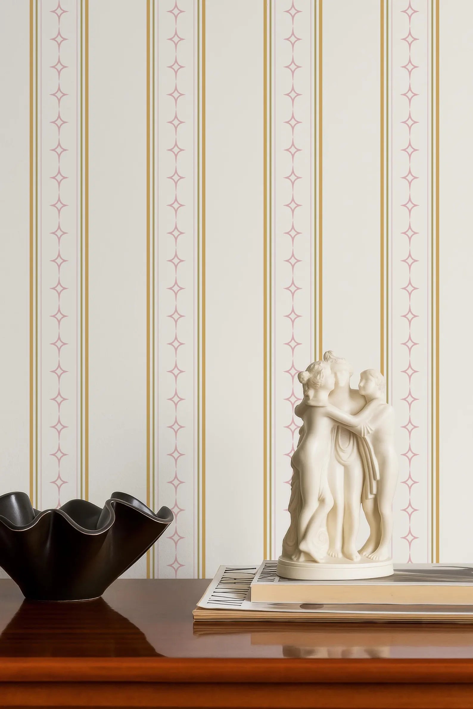 a wooden table with a black bowl, a white figurine, and a stack of books. The table is set against a striped wallpaper with a repeating pattern of pink and gold lines.