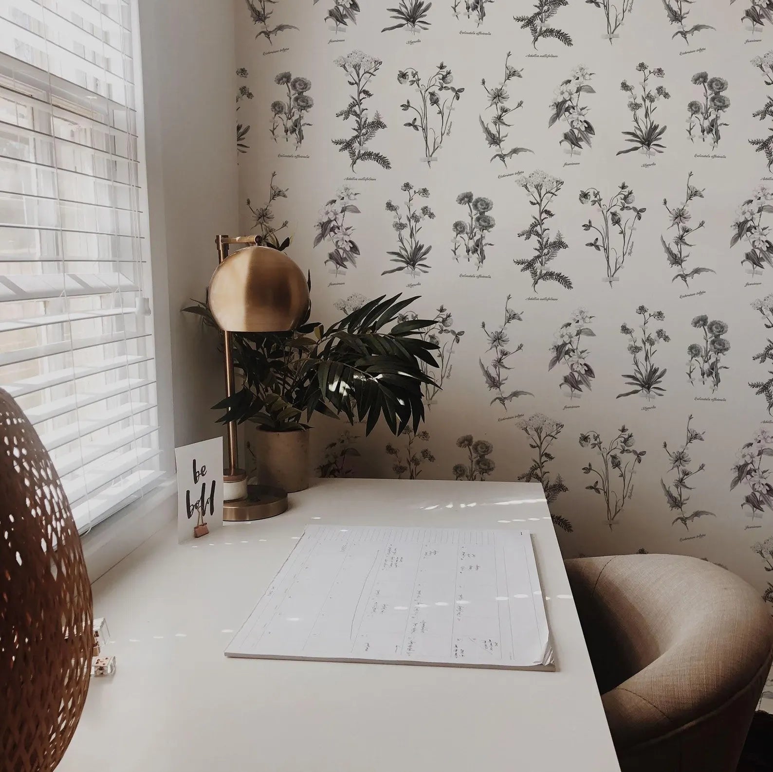 a white desk with a lamp, a plant, and a calendar, set against a wall with a floral wallpaper.