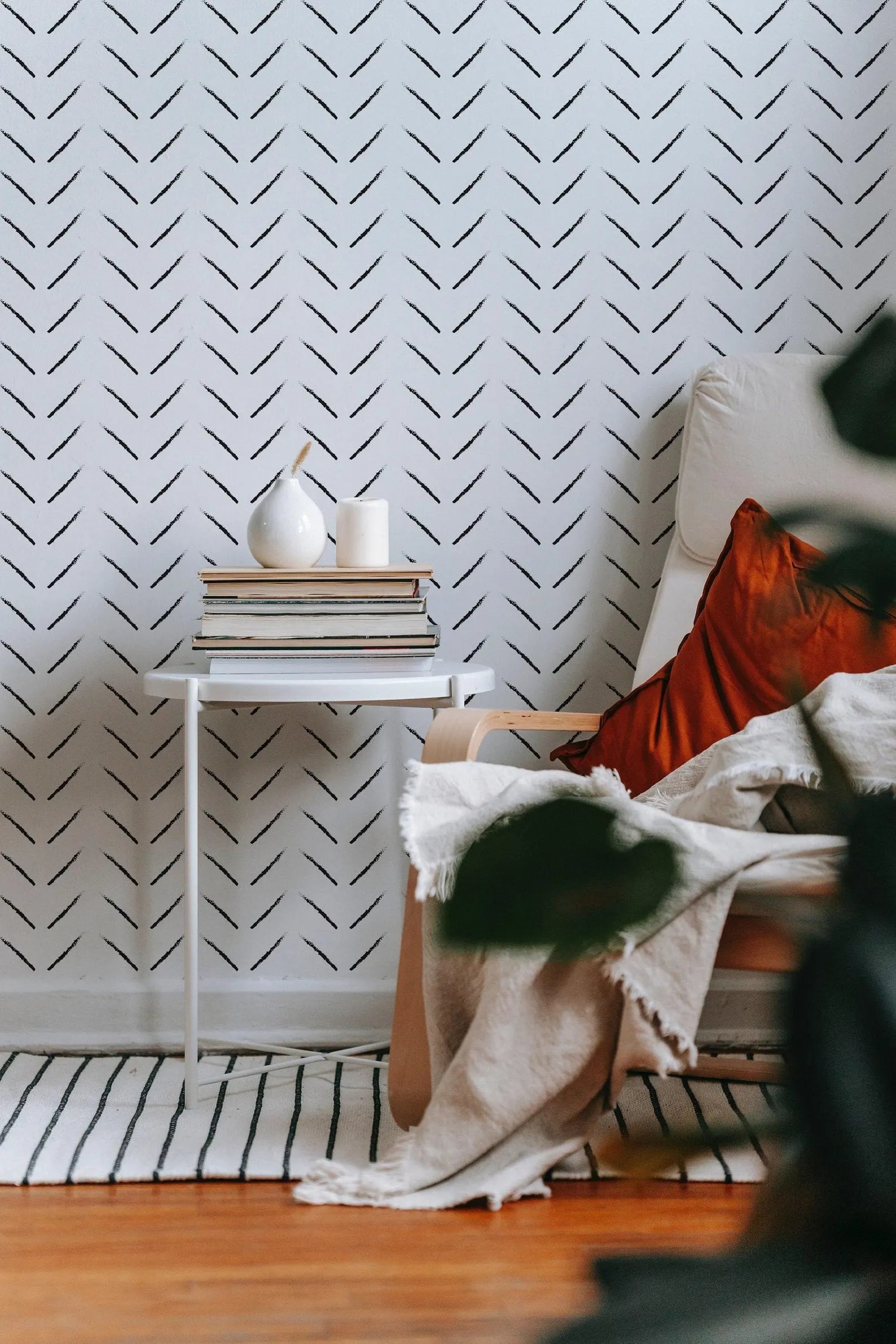 a cozy living space with a white armchair, a white side table, and a stack of books. The wall behind the furniture has a black and white chevron pattern, adding a modern touch to the room. The room is decorated with a white vase, a white candle, and a white bowl, creating a harmonious and inviting atmosphere.