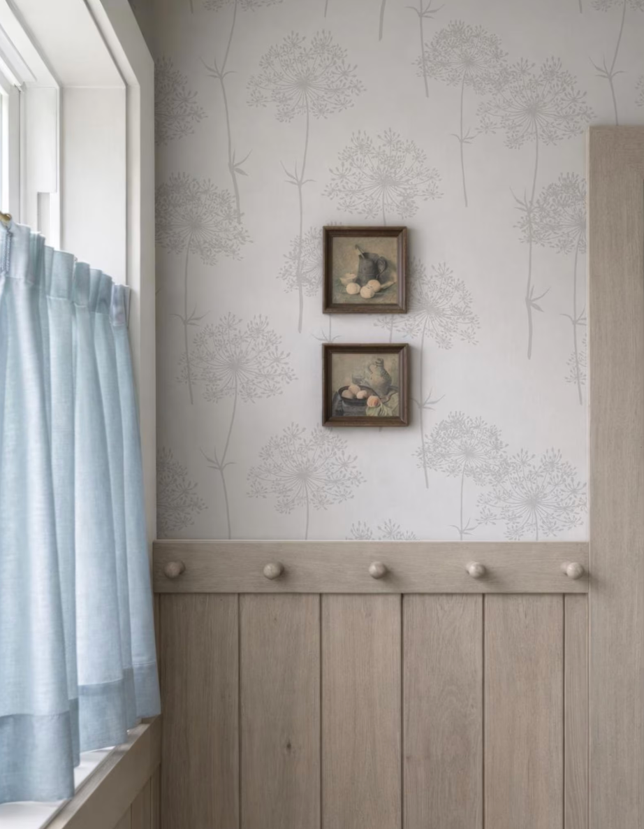 a wall with a gray floral wallpaper, two framed pictures hanging on it, and a window with light blue curtains.