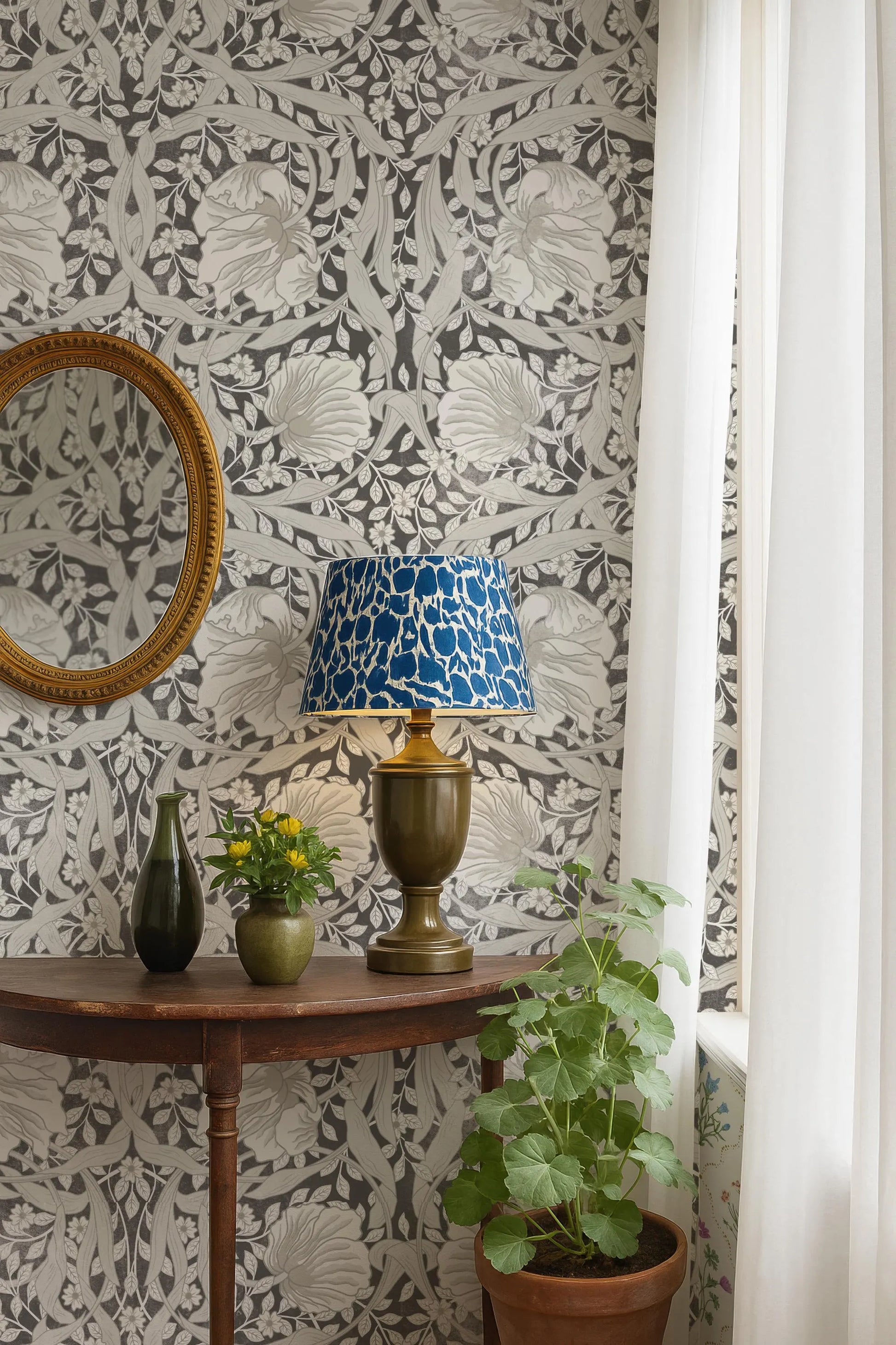 a table with a blue and white patterned lamp shade, a green vase with yellow flowers, and a potted plant. The table is set against a wall with a floral wallpaper pattern.