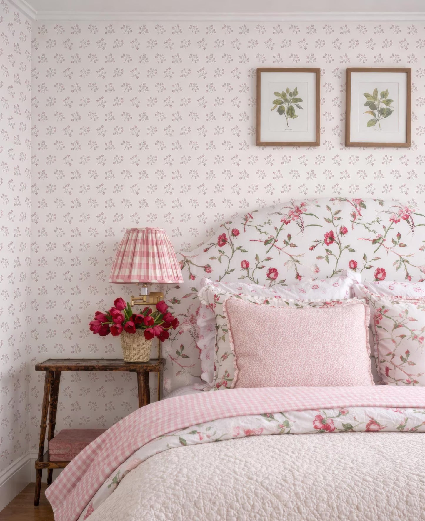 a cozy and charming bedroom with a floral-patterned headboard, a pink and white checkered bedspread, and a wooden nightstand with a lamp and a vase of red flowers. The walls are covered in a floral wallpaper, and there are two framed pictures of flowers above the bed.