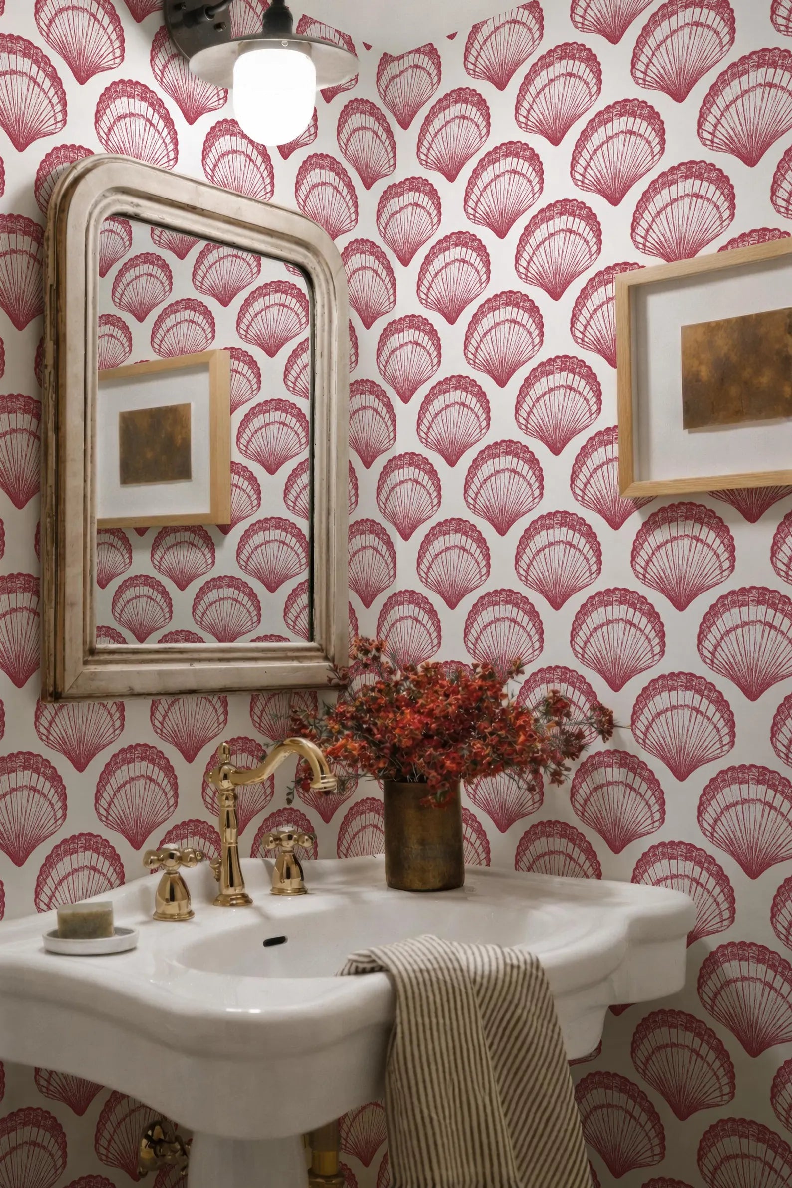 a bathroom with a white pedestal sink, a gold faucet, and a framed picture on the wall. The wall has a red and white patterned wallpaper with a repeating shell design. A vase of red flowers is placed on the sink, and a striped towel is hanging on the side.
