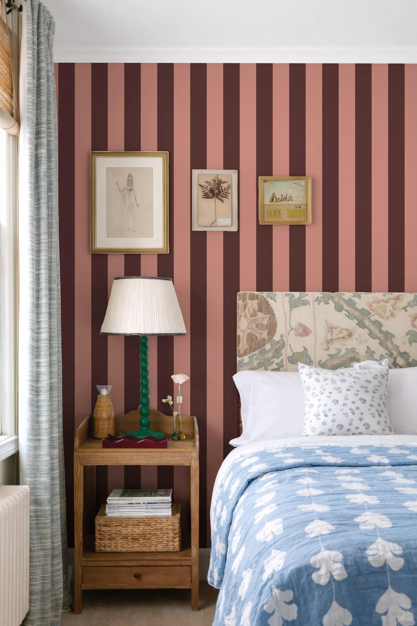 a bedroom with a striped pink and burgundy wall, a floral headboard, and a bed with a blue and white patterned comforter. The room also features a nightstand with a lamp, a picture frame, and a basket of books.
