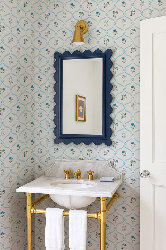 a bathroom with a white sink, a blue scalloped mirror, and a gold faucet. The walls are covered in a floral wallpaper, and there is a white door next to the sink.