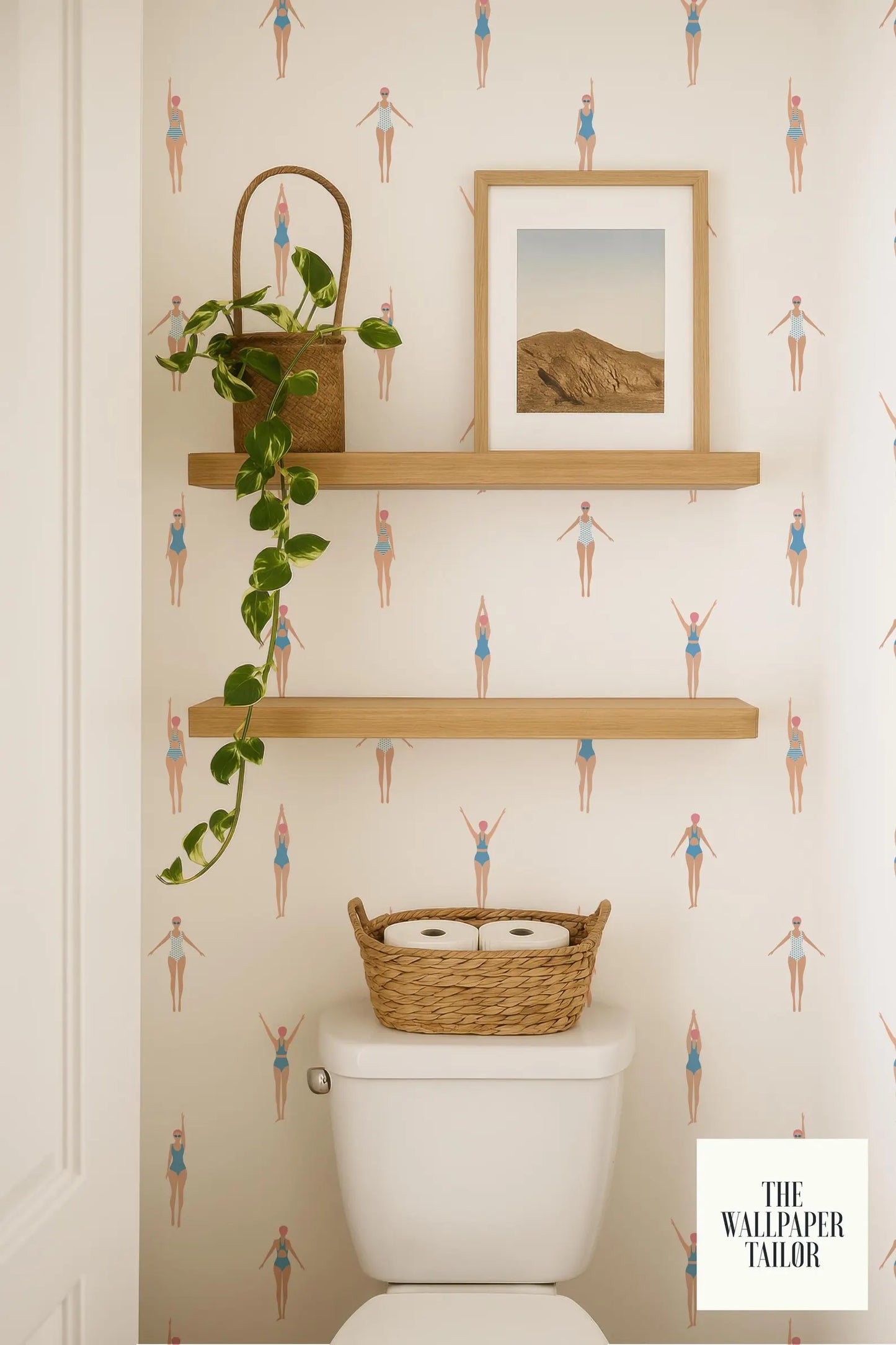 a bathroom with a toilet, a basket of toilet paper, and a framed picture on a shelf. The bathroom has a patterned wallpaper with illustrations of people in swimsuits.