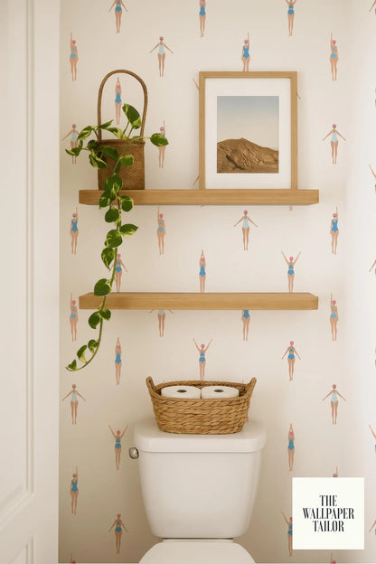 a bathroom with a toilet, a basket of toilet paper, and a framed picture on a shelf. The bathroom has a patterned wallpaper with illustrations of people in swimsuits.