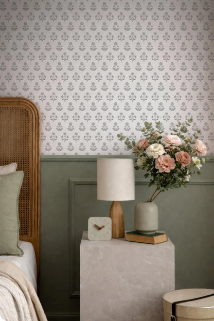 a bedroom setting with a floral wallpaper, a wooden headboard, a white lamp, a vase of pink flowers, and a small table with a clock and a book.