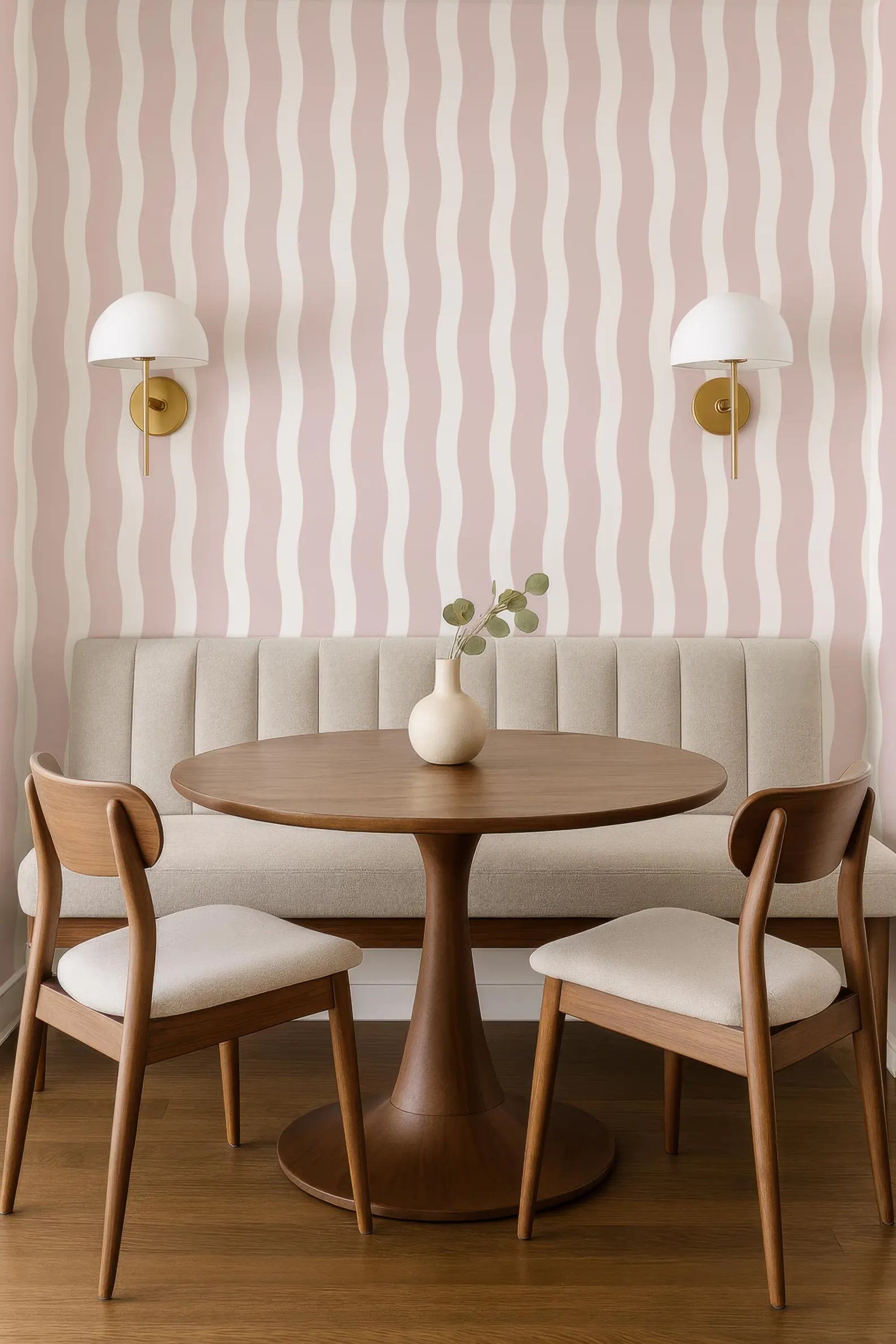 a modern dining area with a round wooden table, two wooden chairs, and a bench. The walls are adorned with pink and white striped wallpaper, and there are two wall sconces on either side of the table. The table is set with a vase of greenery, creating a cozy and inviting atmosphere.