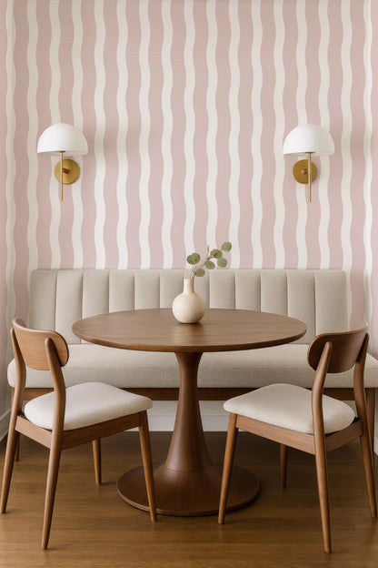 a modern dining area with a round wooden table, two wooden chairs, and a bench. The walls are adorned with pink and white striped wallpaper, and there are two wall sconces on either side of the table. The table is set with a vase of greenery, creating a cozy and inviting atmosphere.