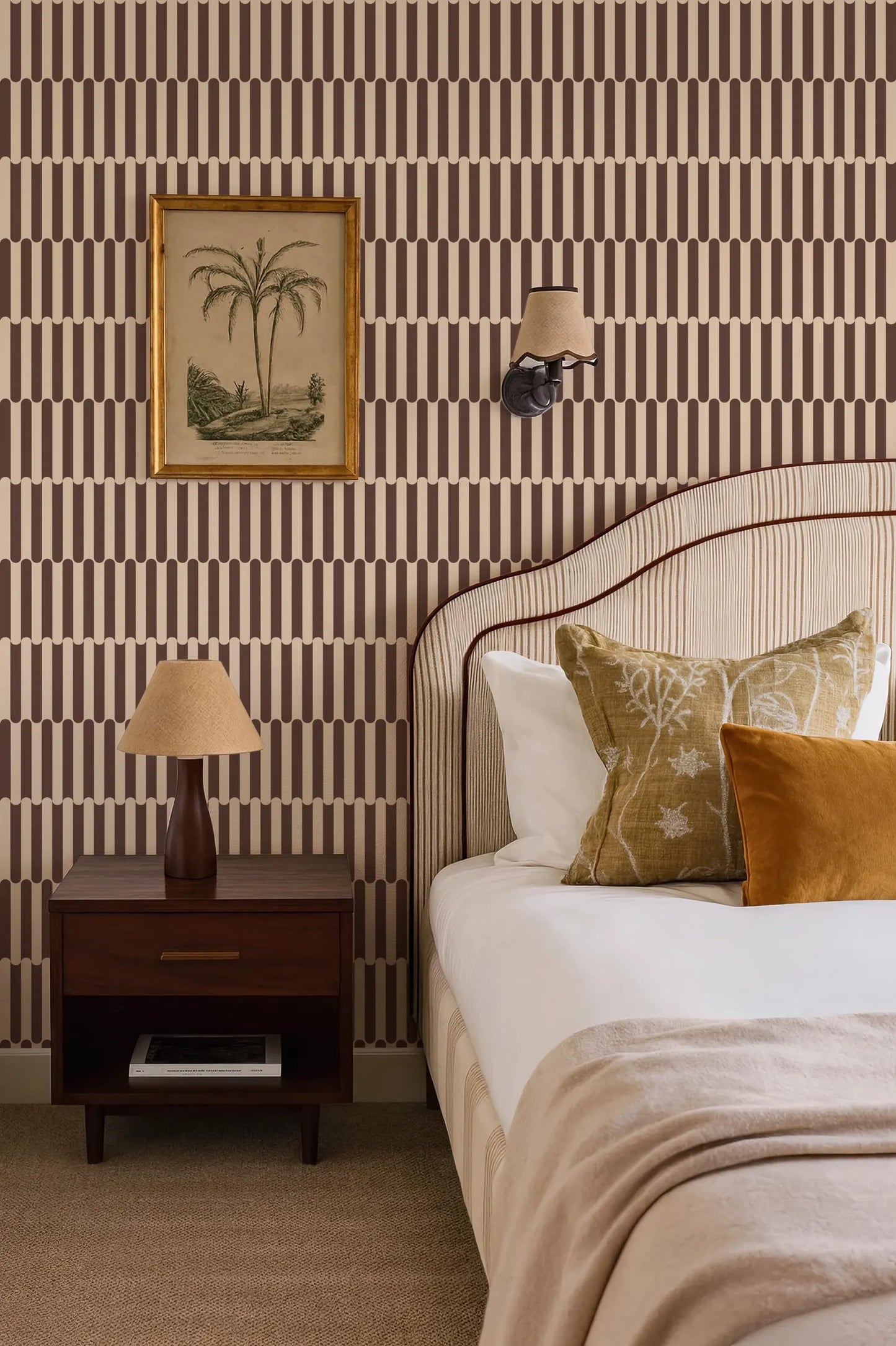 a well-decorated bedroom with a bed, nightstand, and wall art. The bed has a striped headboard and is adorned with decorative pillows. The nightstand has a lamp and a framed picture on its surface. The wall behind the bed is covered in a patterned wallpaper with vertical stripes in various shades of brown and beige.