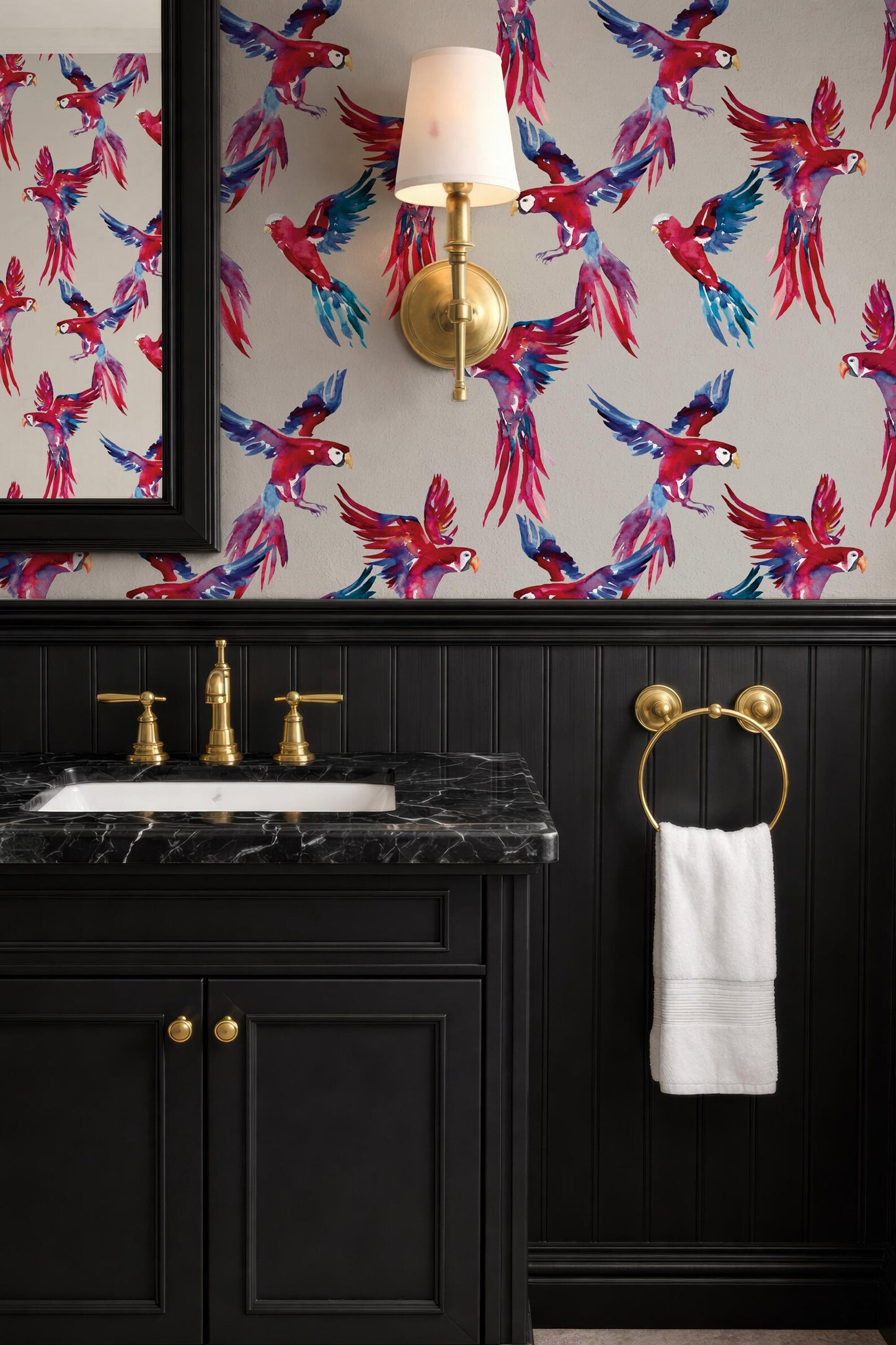 a bathroom with a black vanity, a gold faucet, and a white towel hanging on a gold ring. The walls are covered in a vibrant, colorful wallpaper with a pattern of birds. A gold wall sconce is mounted above the vanity, and a mirror is also present in the bathroom.