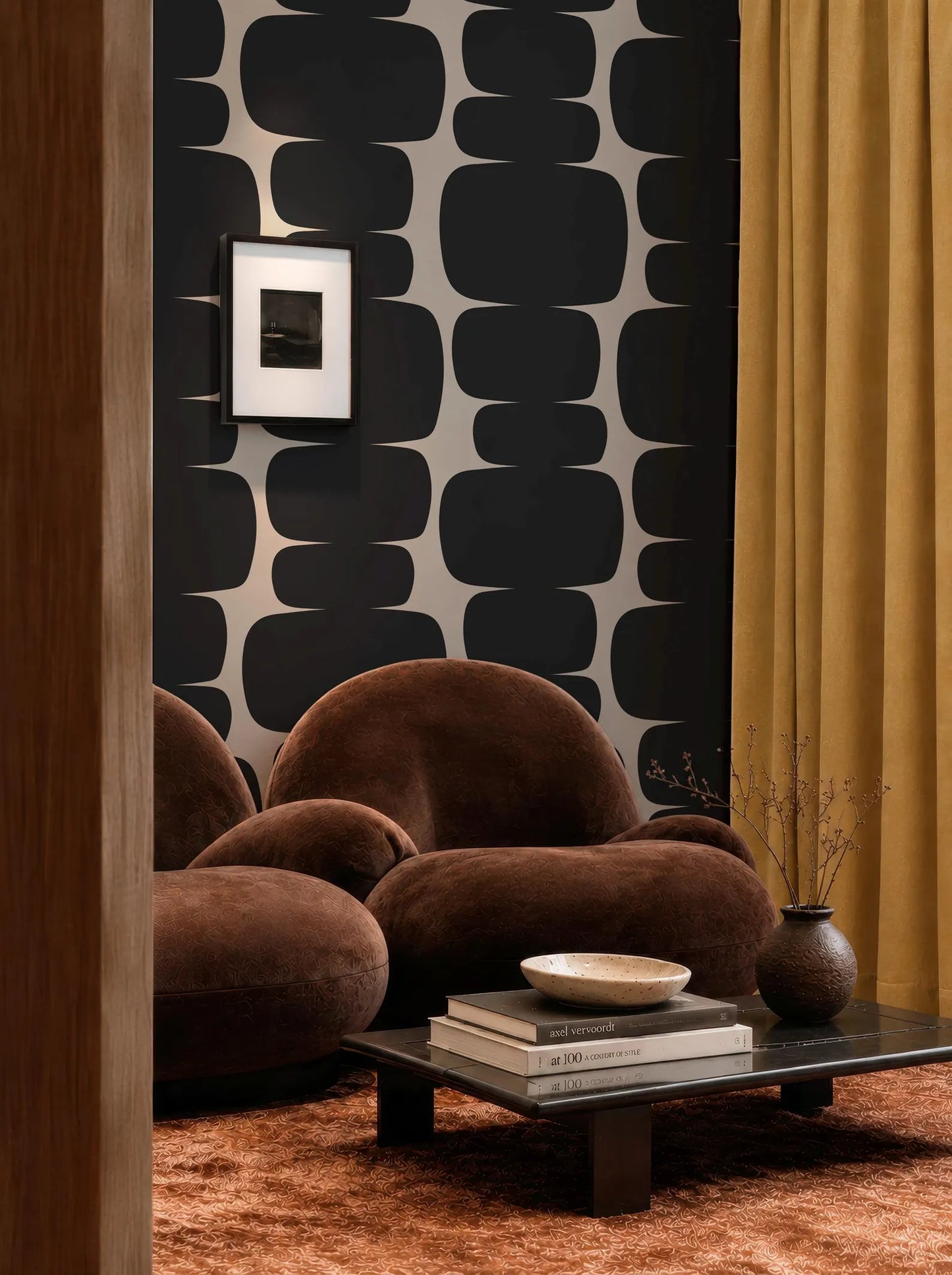 a modern living room with a brown sofa, a coffee table, and a framed picture on the wall. The room has a black and white geometric pattern on the wall, and a yellow curtain. There is also a stack of books on the coffee table.