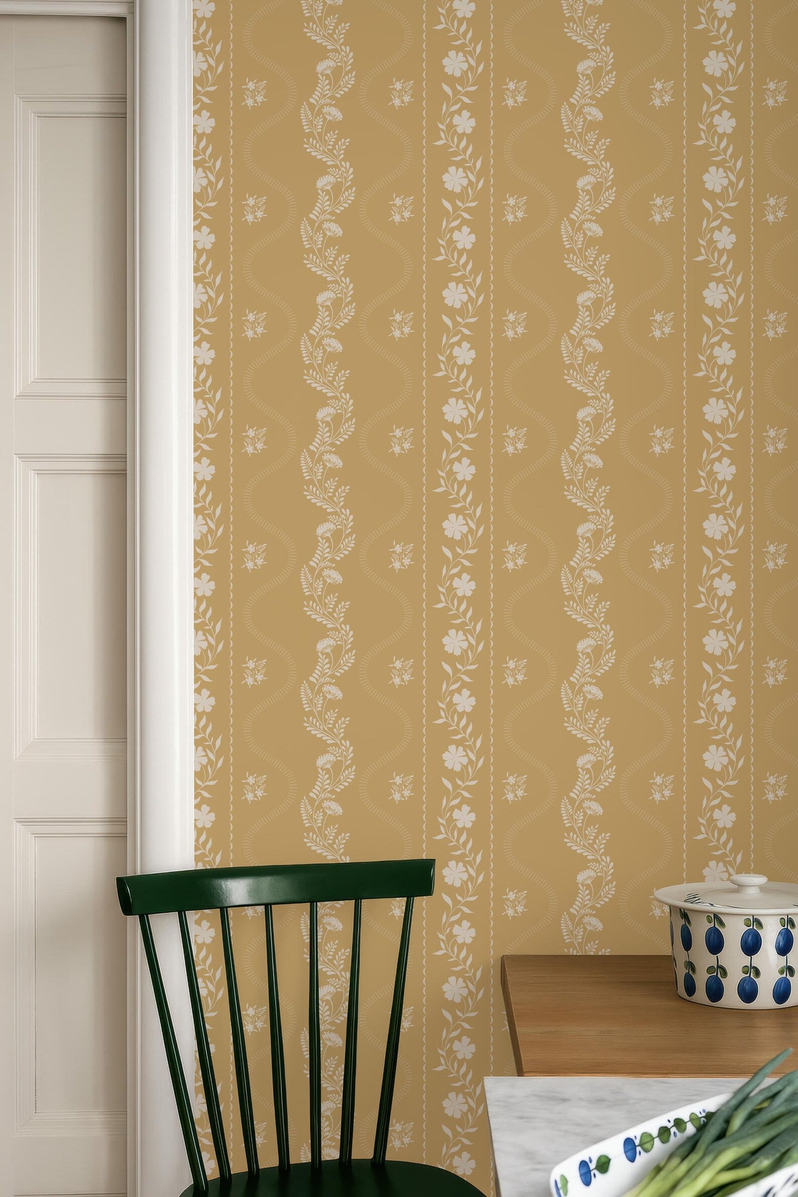 a kitchen interior with a wooden table, a green chair, and a white bowl with blue and green designs on it. The wall behind the table has a patterned wallpaper with vertical lines and floral designs.
