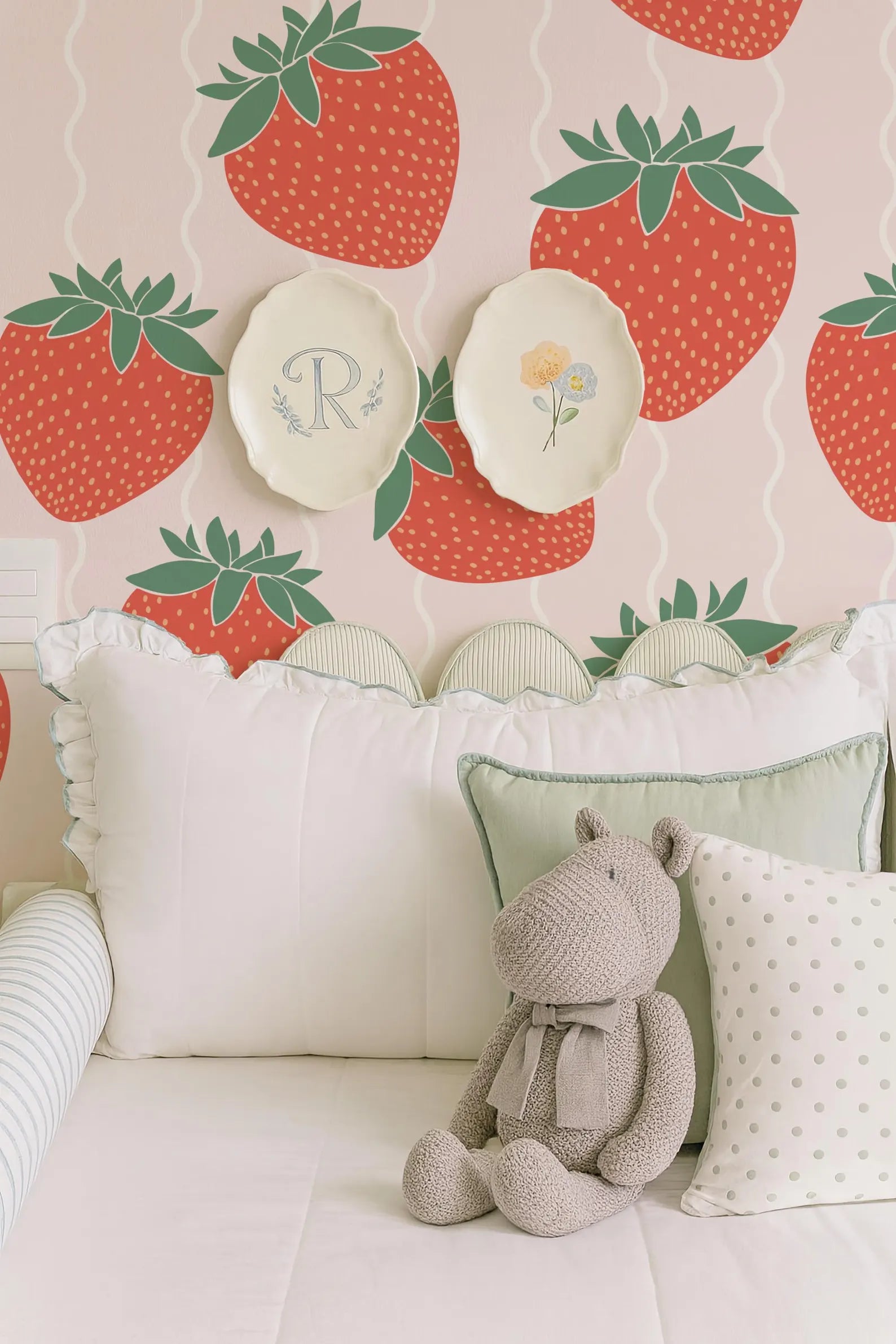 a cozy bedroom scene with a stuffed animal hippo sitting on a bed, surrounded by pillows and a wall with a strawberry wallpaper.