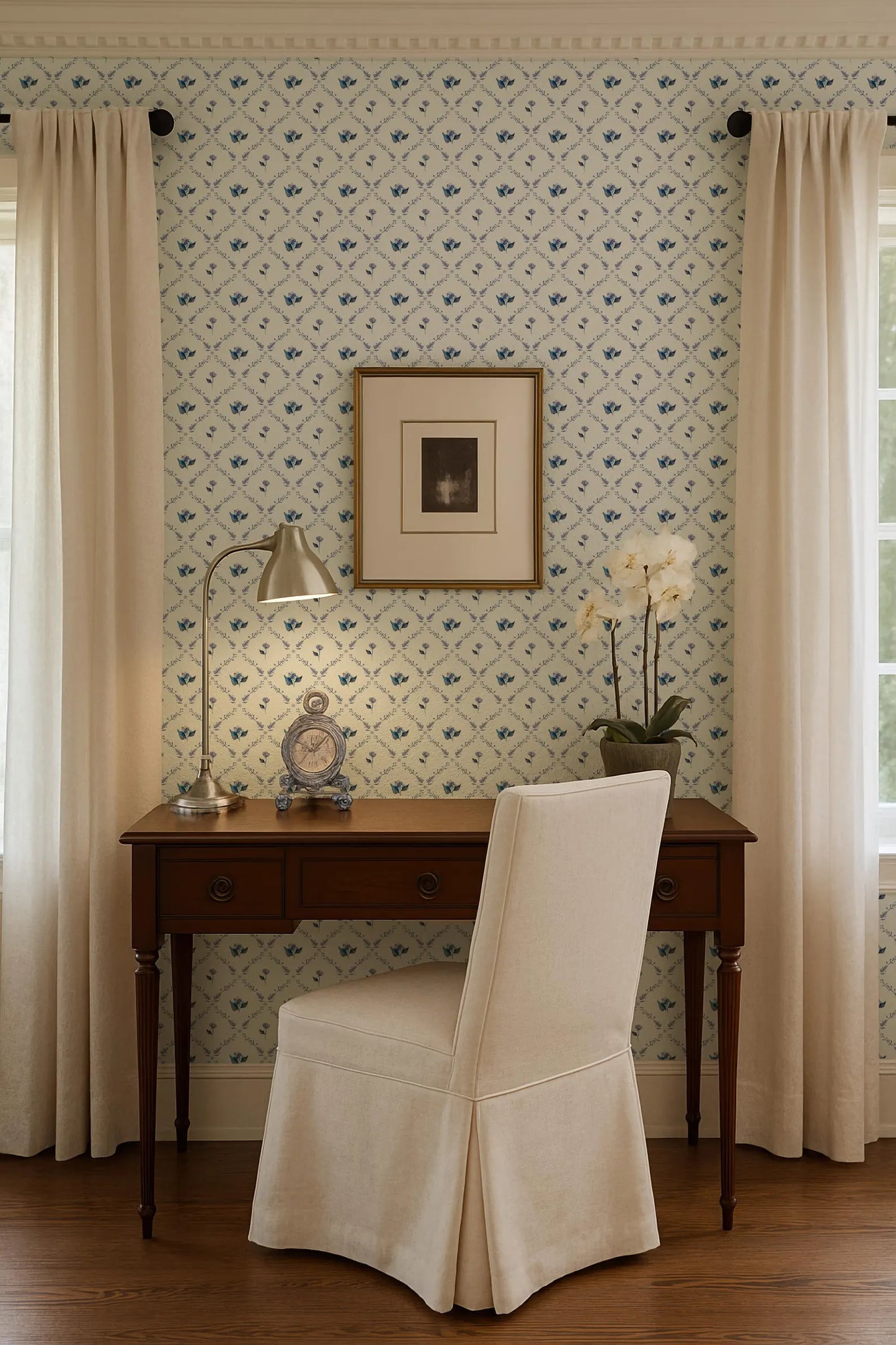 a cozy corner of a room with a wooden desk, a white chair, and a framed picture on the wall. The room has a patterned wallpaper and a window with curtains.