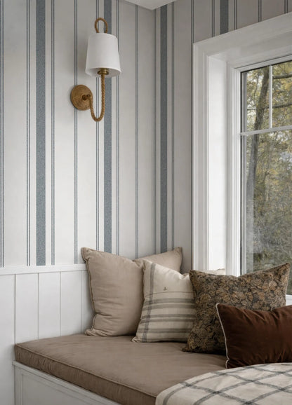 a cozy corner of a room with a window, a white wall, and a striped wallpaper. There is a bed with a brown and white striped pillow and a white and gray striped pillow. A wall sconce is mounted on the wall above the bed.
