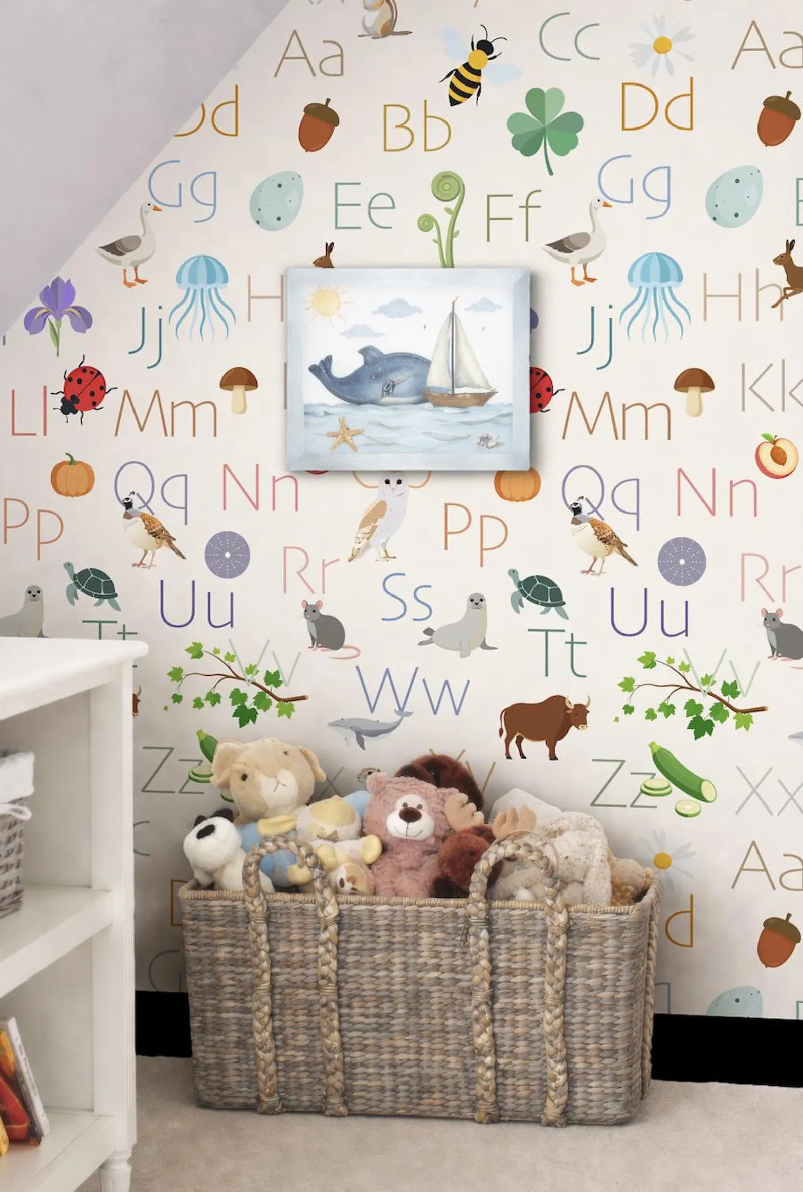 A wall with a colorful alphabet wallpapered wall, featuring various animal and letter illustrations. In the foreground, there is a wicker basket filled with stuffed animals, and a framed picture of a whale is hanging on the wall above the basket.
