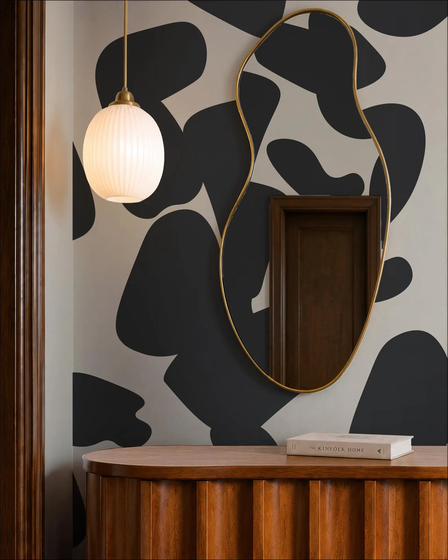 a modern interior design with a black and white abstract pattern on the wall behind a wooden dresser. A round mirror with a gold frame hangs above the dresser, and a white pendant light hangs from the ceiling. On the dresser, there is a book titled "The Kinfolk Home".