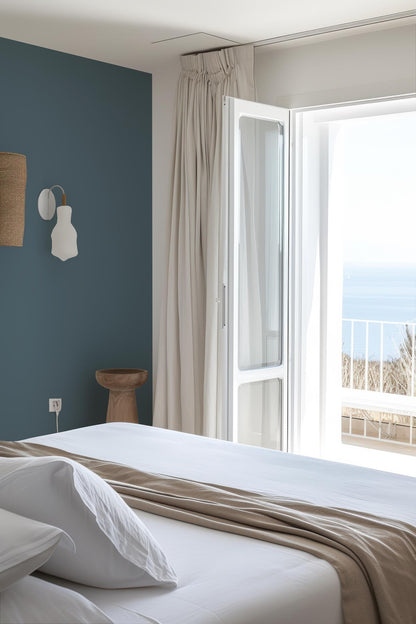 a serene bedroom with a large bed, a wooden stool, and a view of the ocean through a large window.