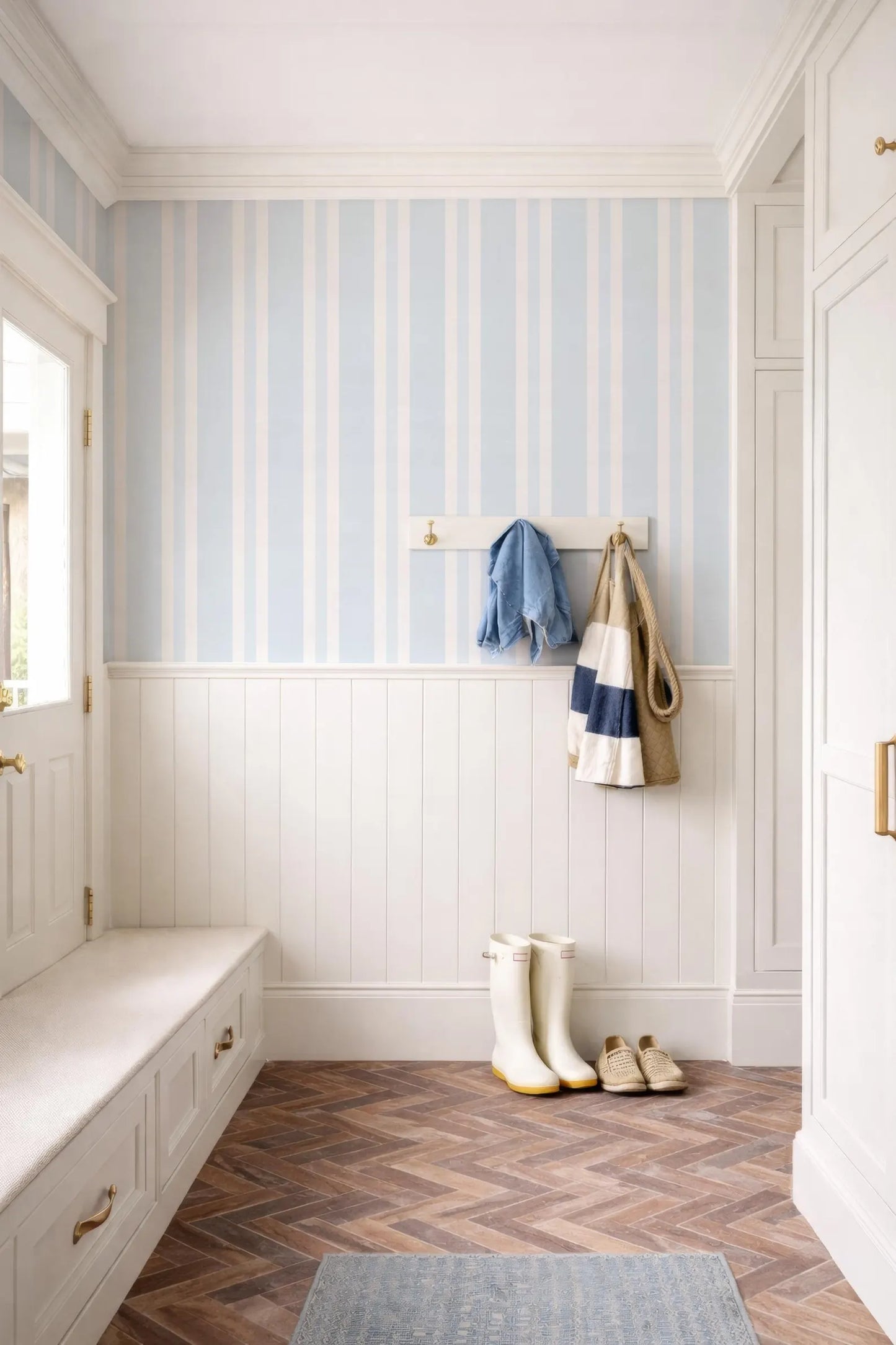 a well-organized entryway with a white bench, a coat rack with a blue and white striped towel, and a pair of white rain boots. The floor is made of wood, and the walls are painted in a light blue color with white stripes.