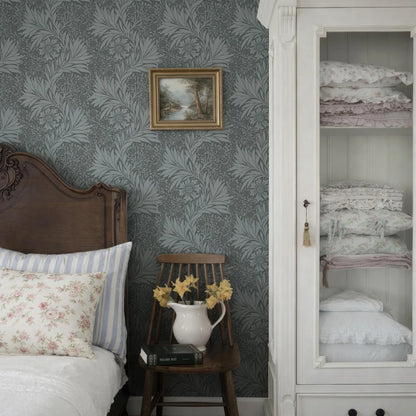 a cozy bedroom scene with a wooden headboard, a white bedspread, and a wooden chair with a vase of yellow flowers. The walls are covered in a gray and white patterned wallpaper, and there is a framed picture hanging on the wall.