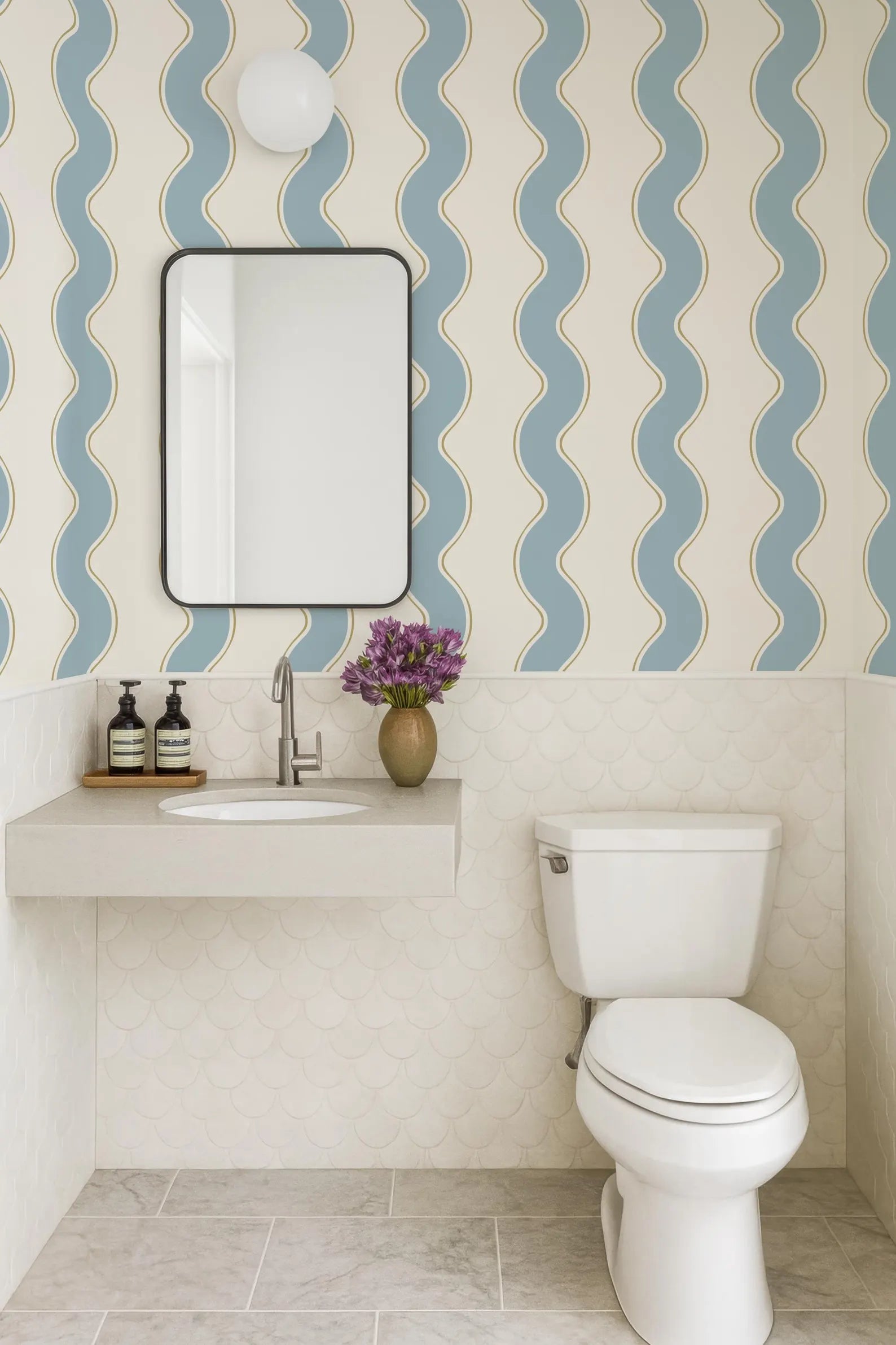 a bathroom with a white toilet, a sink with a mirror above it, and a vase of purple flowers on the counter. The walls are covered in a patterned wallpaper with blue and white stripes.