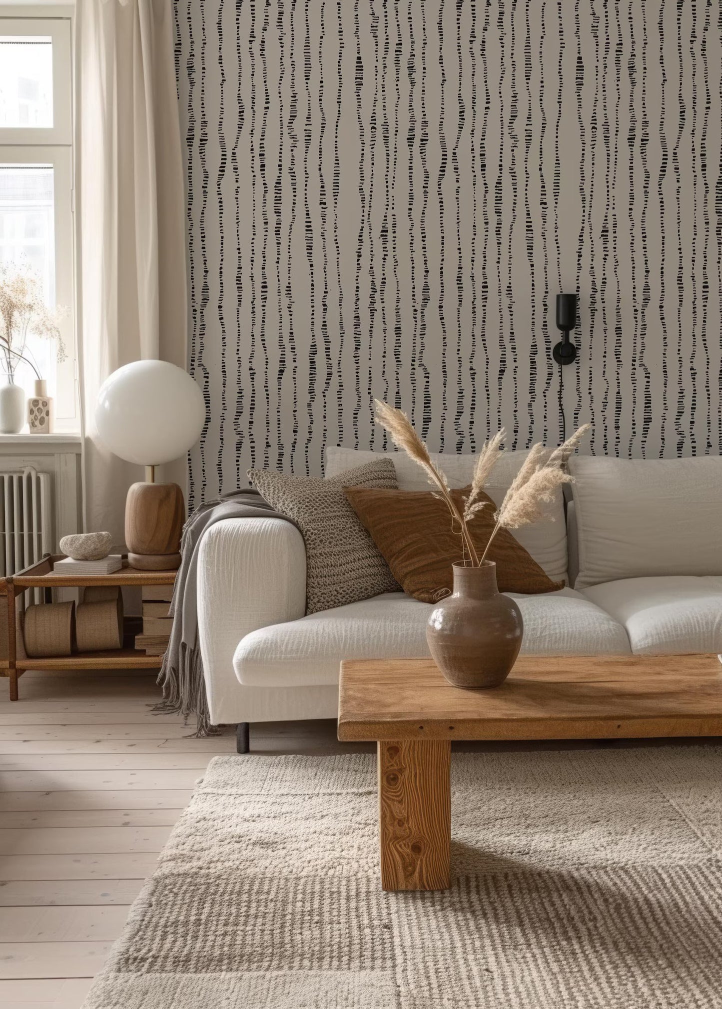 a cozy living room with a white sofa, a wooden coffee table, and a patterned rug. The room has a neutral color palette with a black and white striped wallpaper, and a wooden side table with a lamp and a vase of dried flowers.