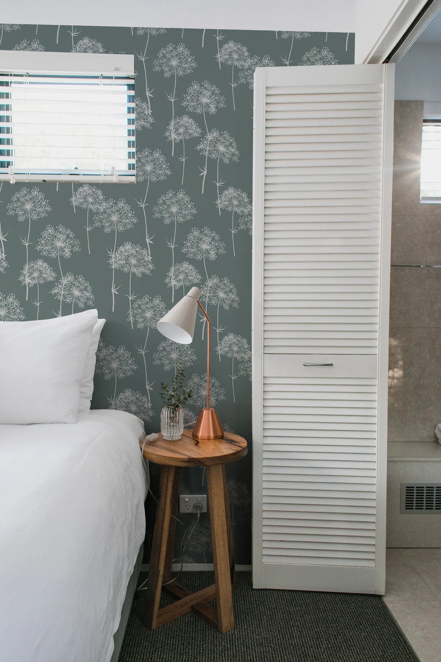 a cozy bedroom with a bed, a nightstand, and a white door with a window. The room has a green and white patterned wallpaper, and the bed is neatly made with white linens.