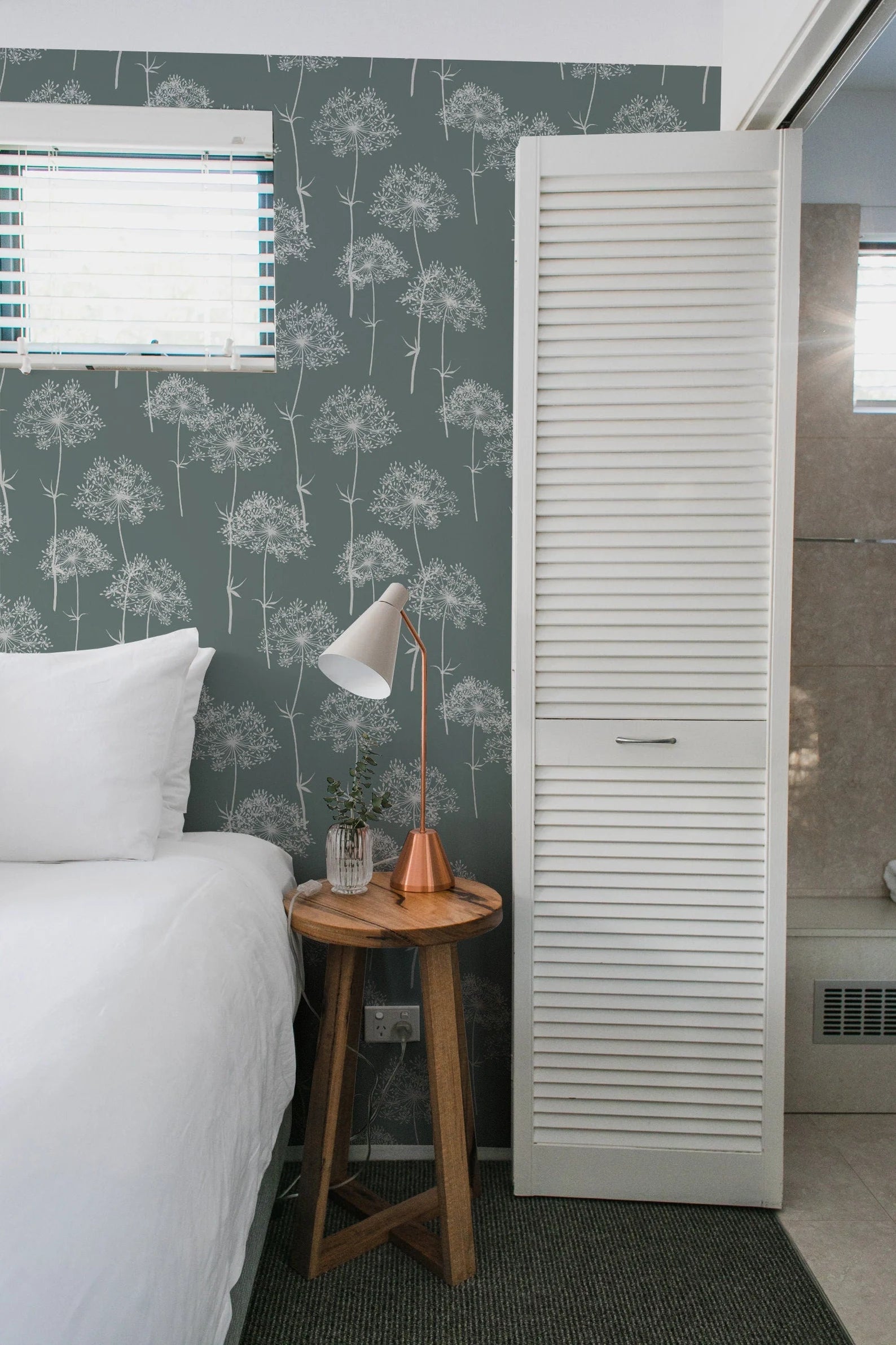 a cozy bedroom with a bed, a nightstand, and a white door with a window. The room has a green and white patterned wallpaper, and the bed is neatly made with white linens.