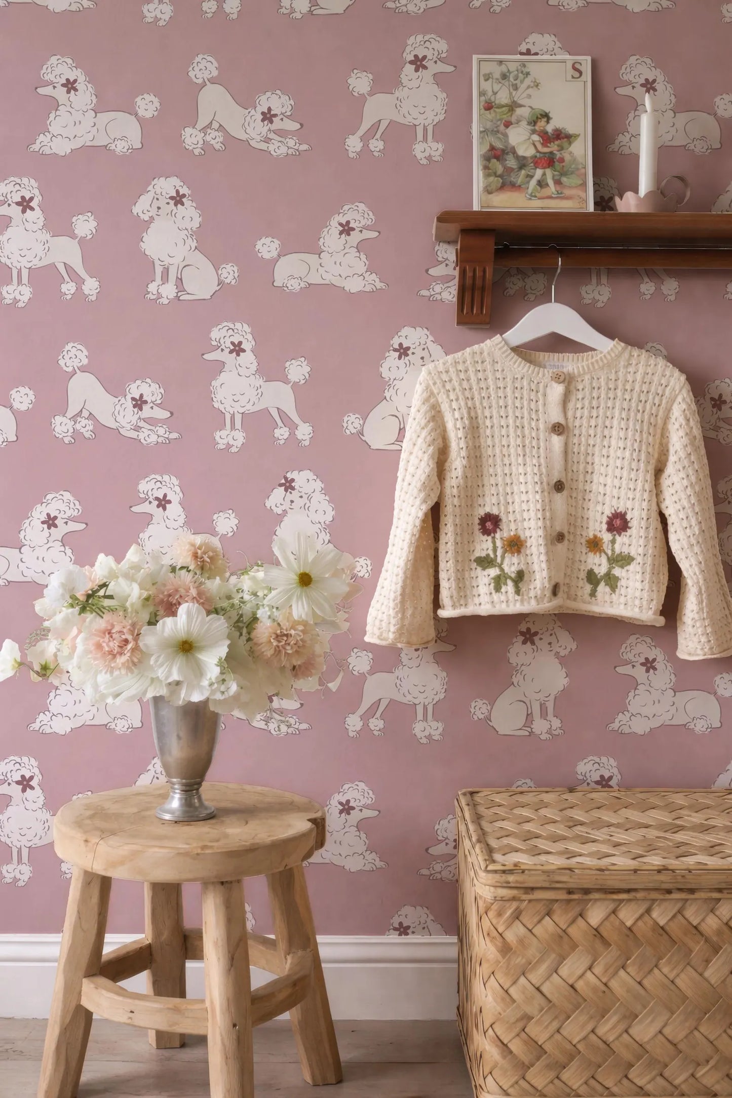 a pink wall with a pattern of white poodles, a wooden stool with a vase of flowers, a wooden shelf with a sweater, and a woven basket.