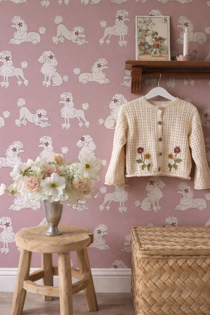 a pink wall with a pattern of white poodles, a wooden stool with a vase of flowers, a wooden shelf with a sweater, and a woven basket.