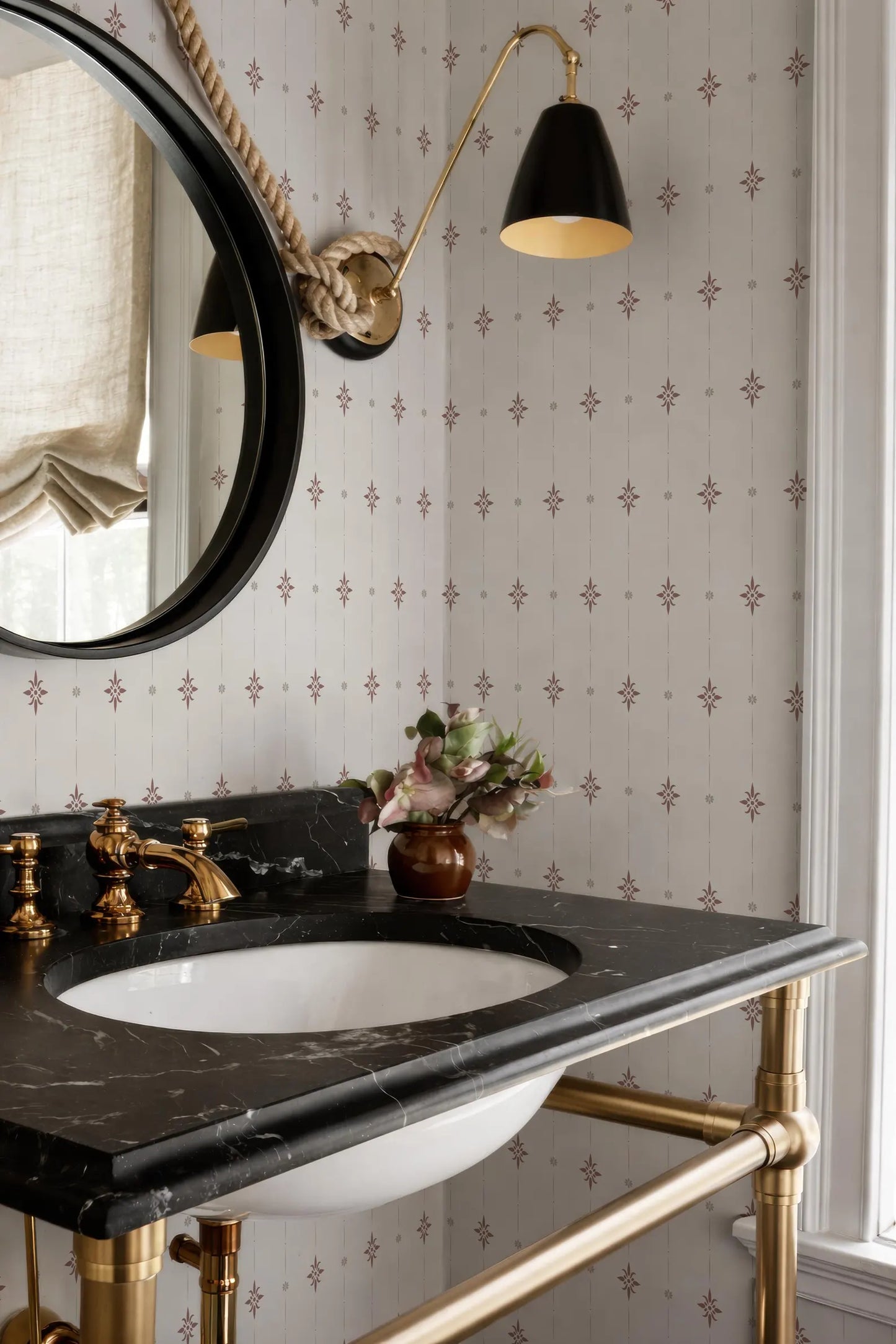 a bathroom with a black marble sink, a round mirror, and a black and gold light fixture above the mirror. The walls are decorated with a patterned wallpaper, and there is a vase of flowers on the countertop.