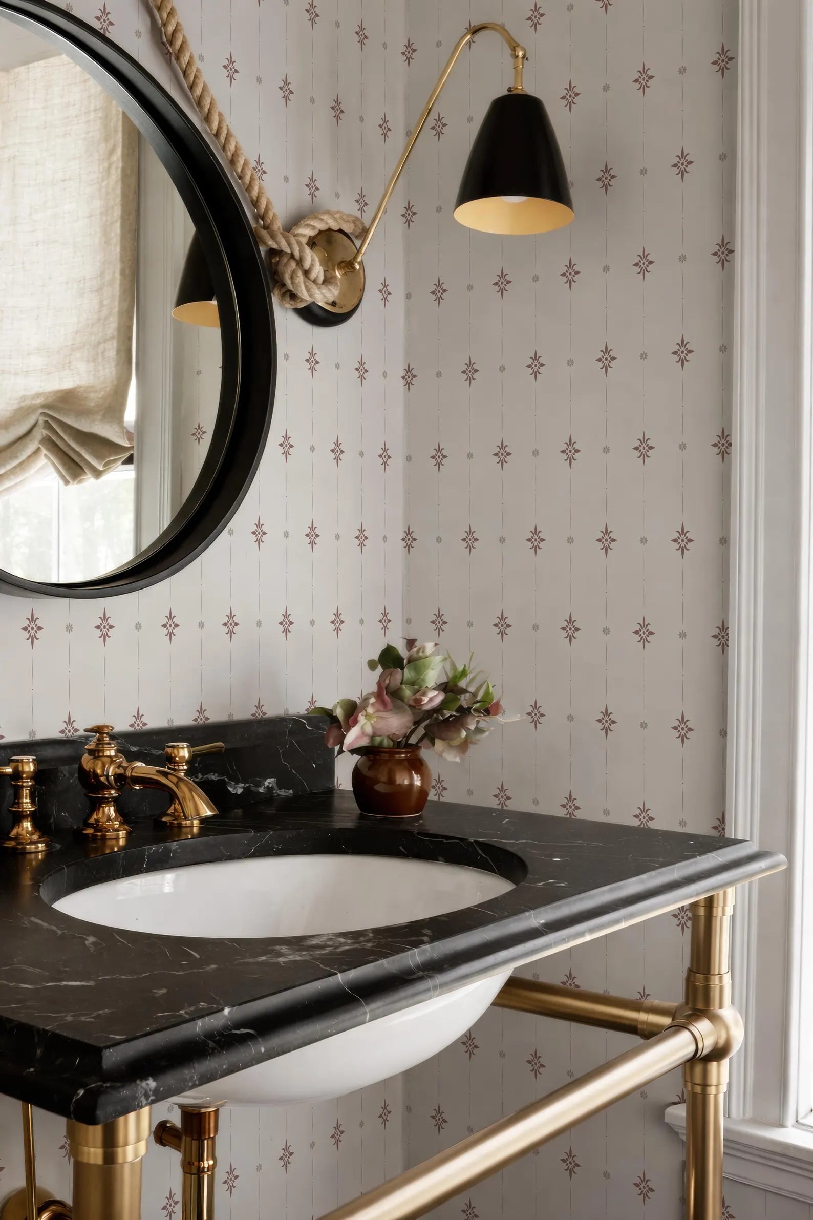 a bathroom with a black marble sink, a round mirror, and a black and gold light fixture above the mirror. The walls are decorated with a patterned wallpaper, and there is a vase of flowers on the countertop.