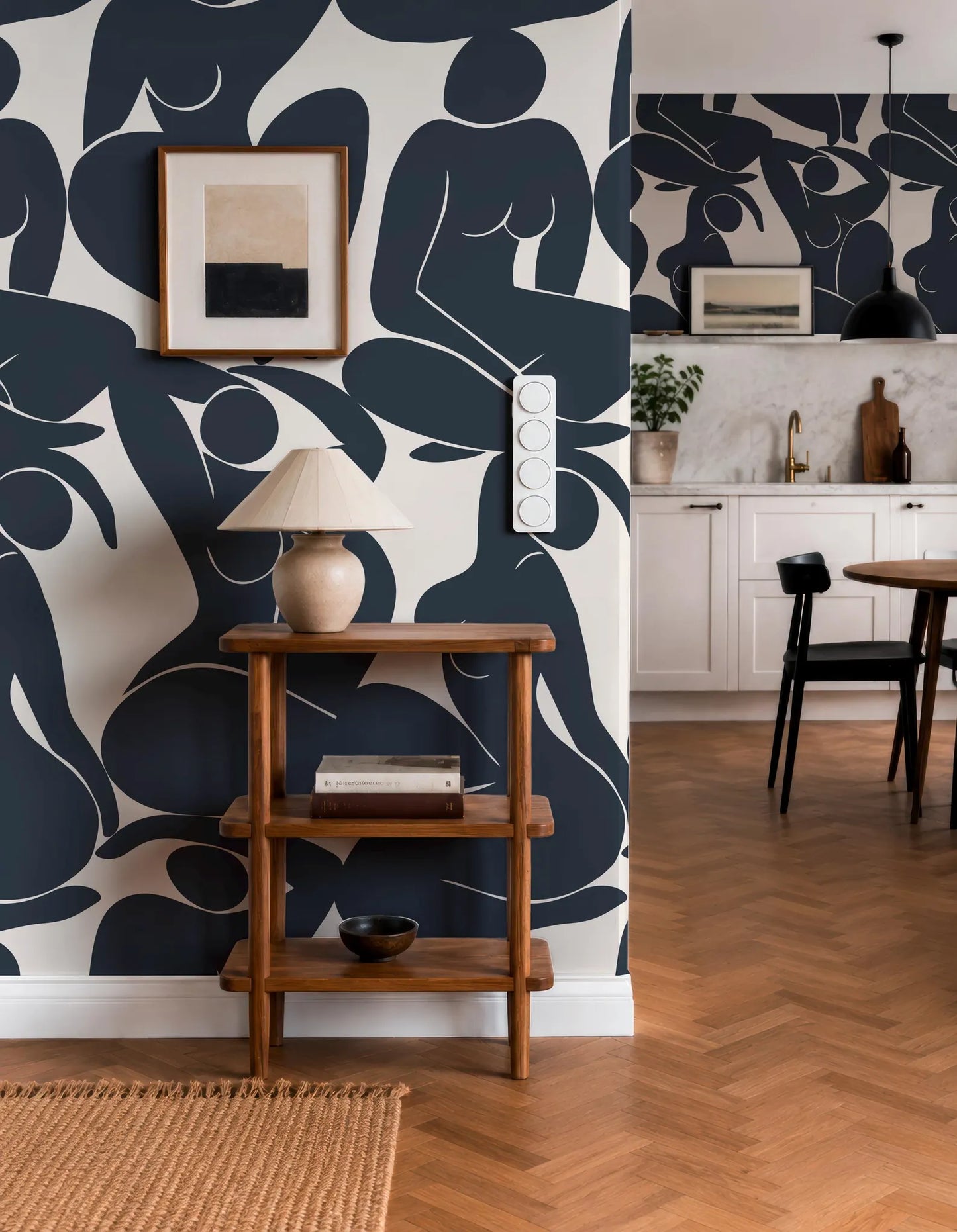 a modern and stylish interior design with a bold black and white abstract wallpaper pattern on the wall. The room includes a wooden side table with a lamp, a framed picture, and a small plant. The dining area has a wooden table and chairs, and the kitchen area has white cabinets and a sink.