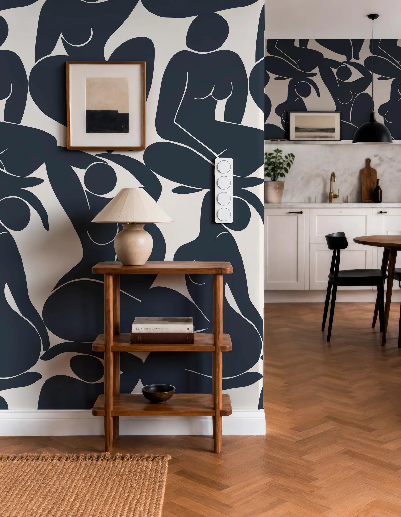 a modern and stylish interior design with a bold black and white abstract wallpaper pattern on the wall. The room includes a wooden side table with a lamp, a framed picture, and a small plant. The dining area has a wooden table and chairs, and the kitchen area has white cabinets and a sink.