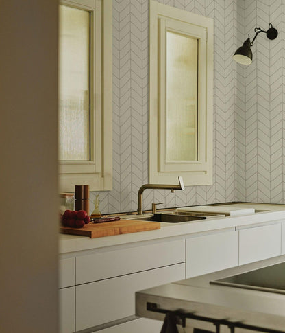 a modern kitchen with a white countertop, a sink, and a wooden cutting board. The kitchen features a chevron patterned wall, and there is a window above the sink.