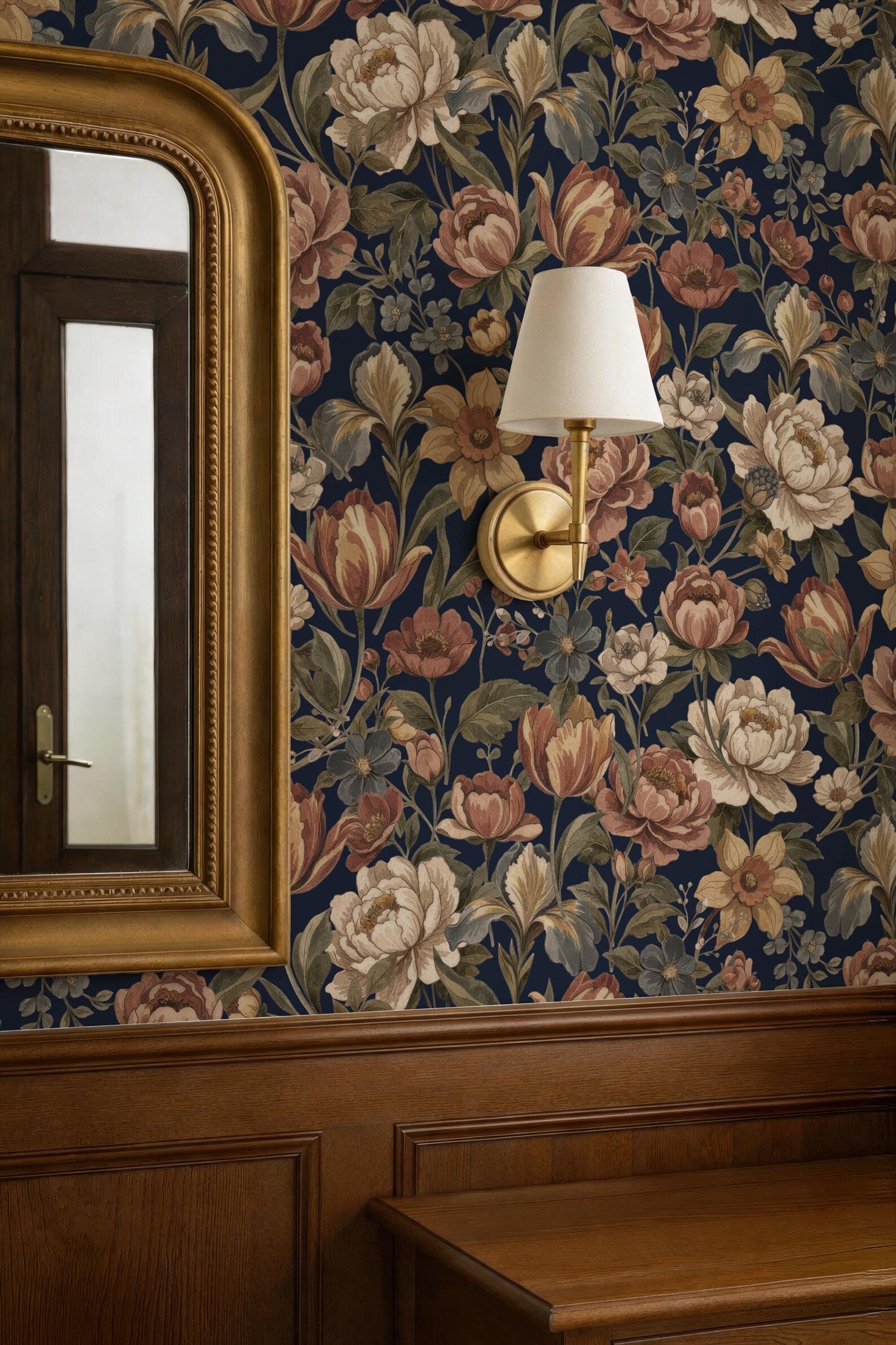 a room with a floral wallpaper, a gold-framed mirror, and a wall-mounted lamp.