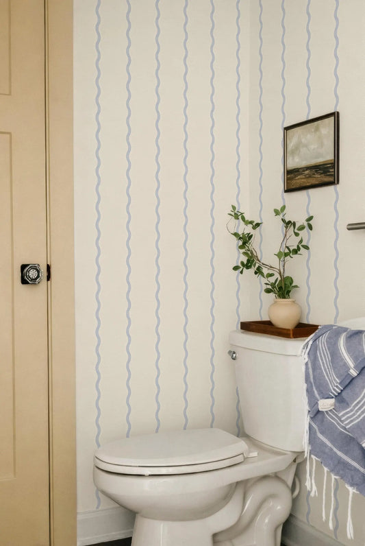a bathroom with a white toilet, a framed picture on the wall, and a potted plant on the tank. The walls are decorated with blue and white striped wallpaper, and the overall color scheme is neutral.
