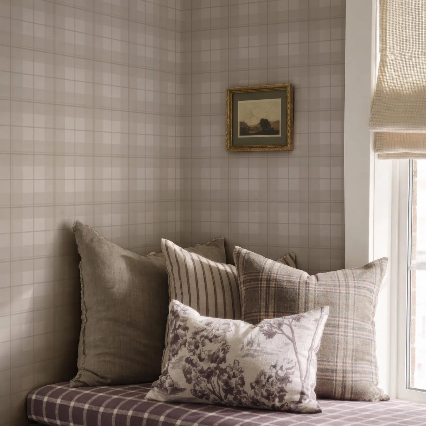 a cozy corner of a room with a plaid patterned couch, a framed landscape painting on the wall, and a window with a beige curtain.