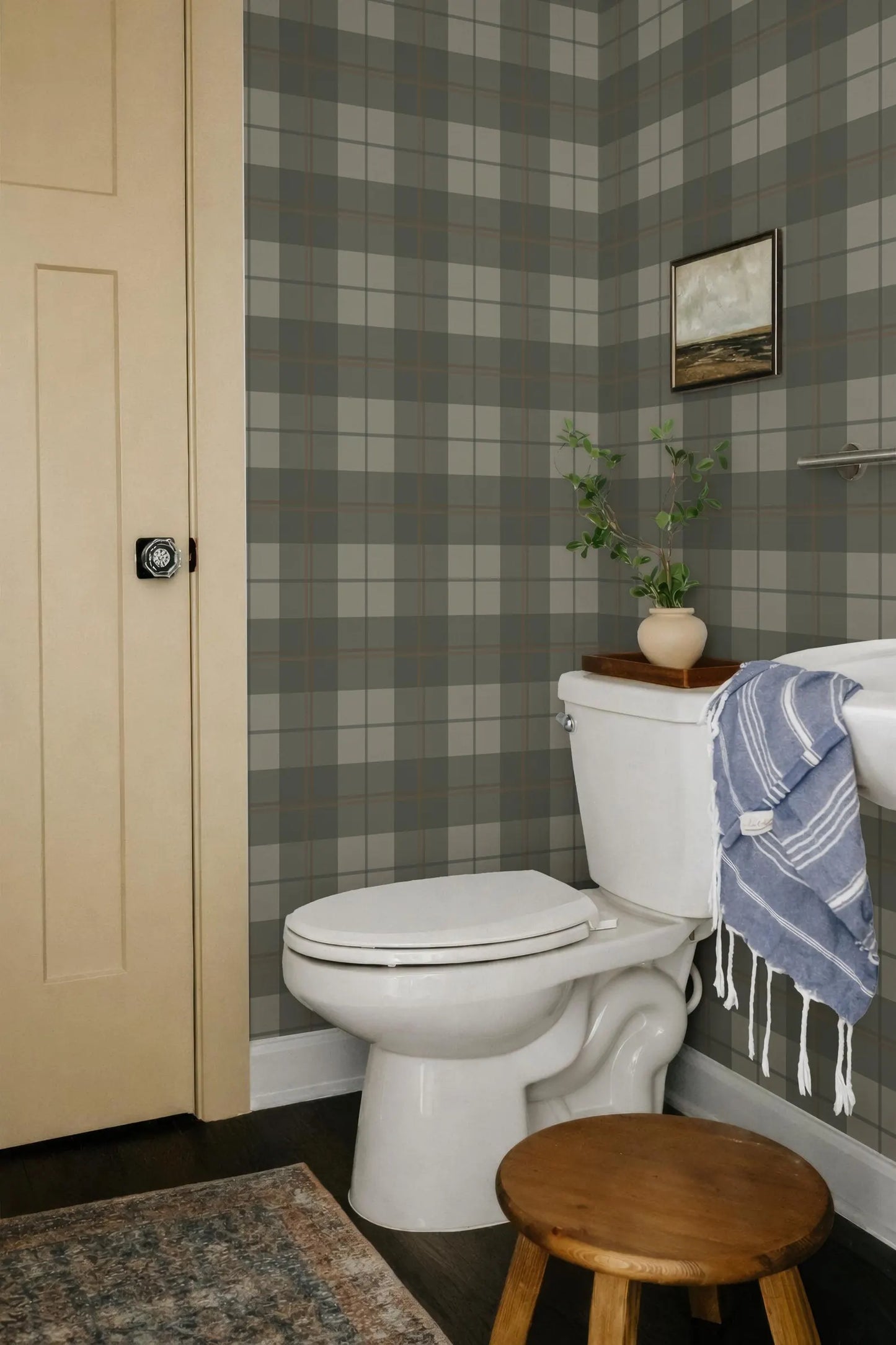 a bathroom with a white toilet, a wooden stool, and a blue and white striped towel hanging on a rack. The walls are covered in a checkered pattern, and there is a painting hanging above the toilet.
