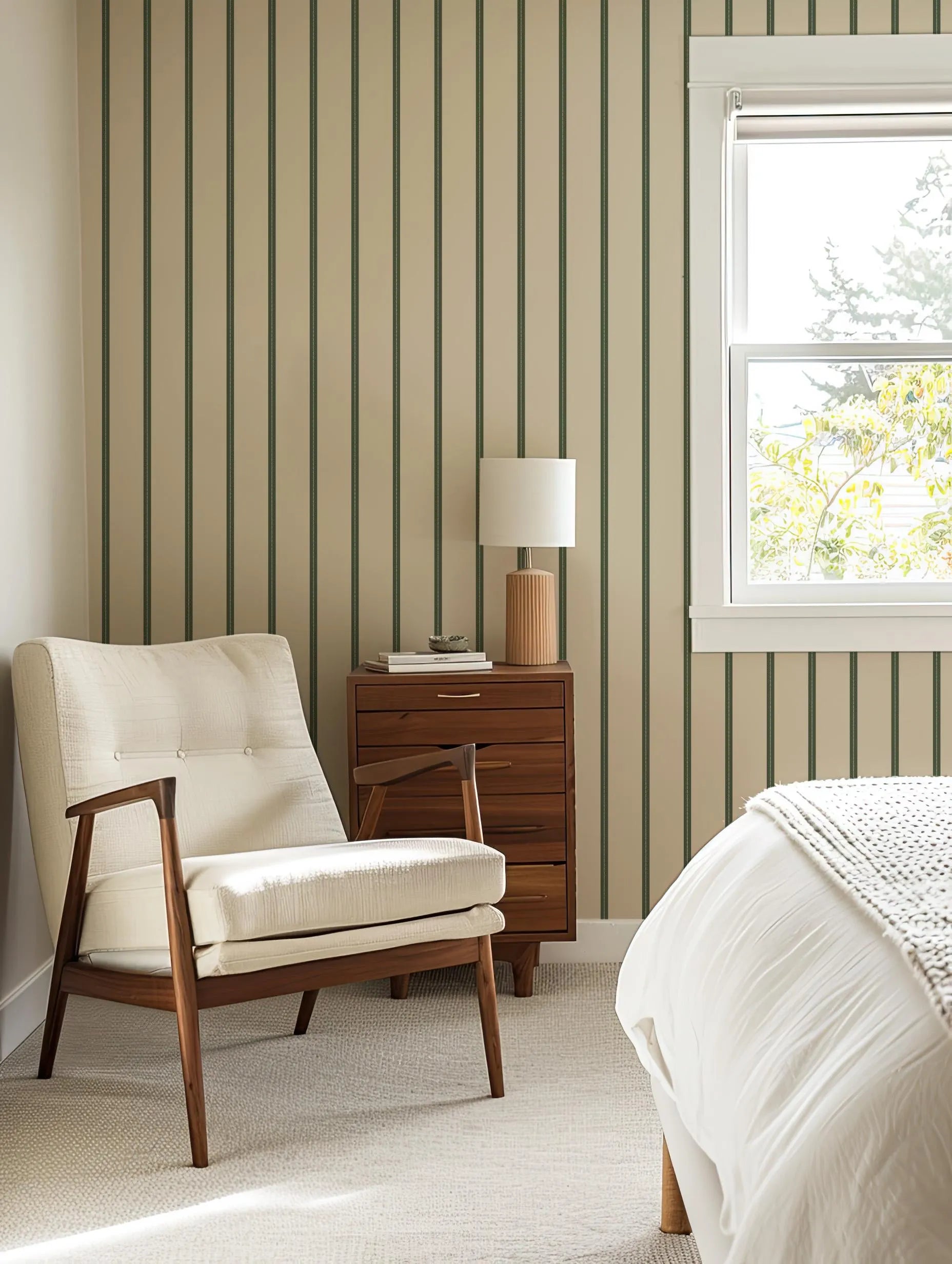 a cozy bedroom with a white armchair, a wooden side table with a lamp, and a window overlooking a tree outside.
