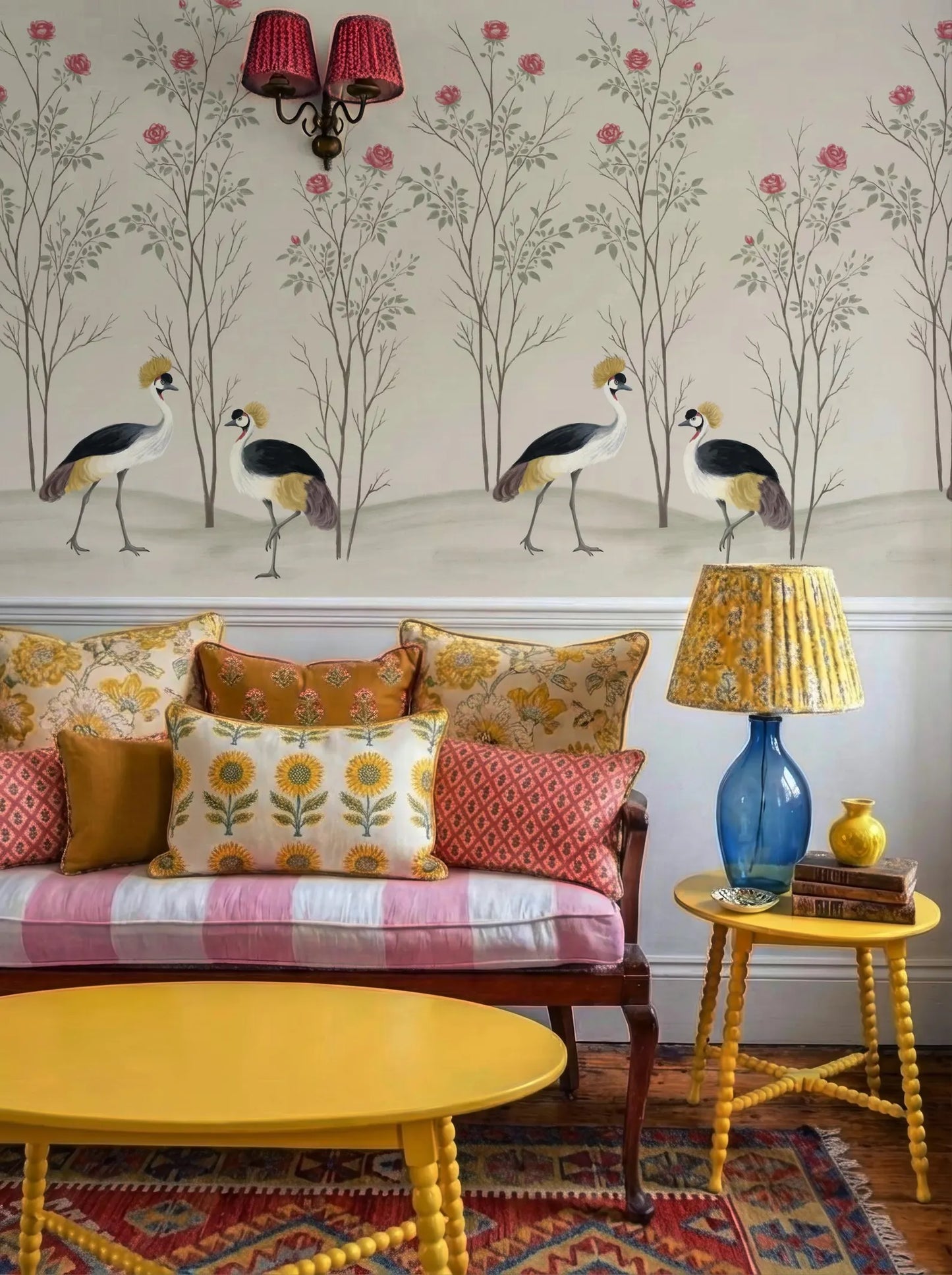 a living room with a yellow sofa adorned with colorful pillows, a yellow side table with a lamp, and a wallpapered wall with a floral design.