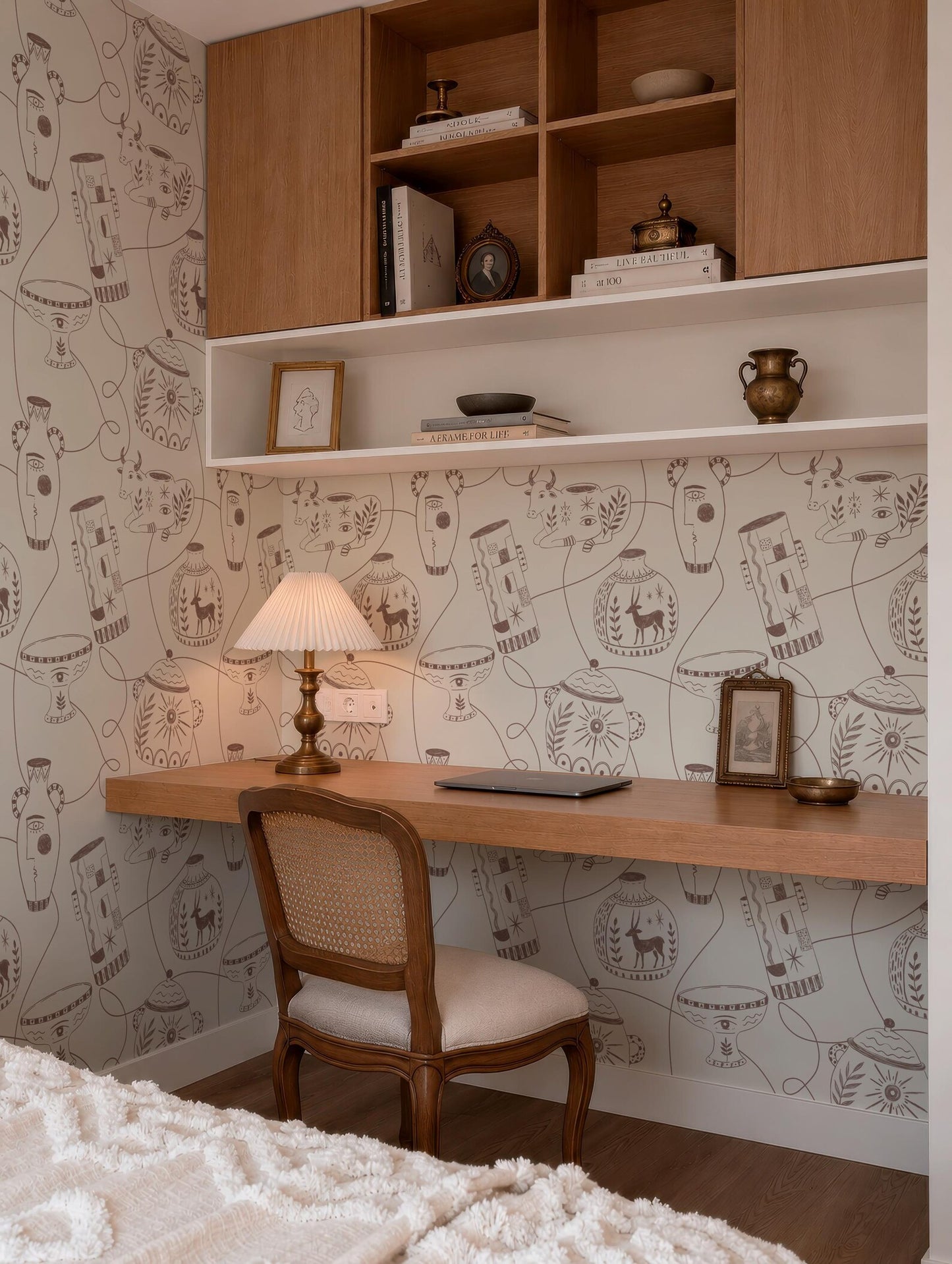a cozy and well-organized home office space with a wooden desk, a comfortable chair, and a bookshelf above it. The wall behind the desk is adorned with a patterned wallpaper, and the room has a warm and inviting atmosphere.