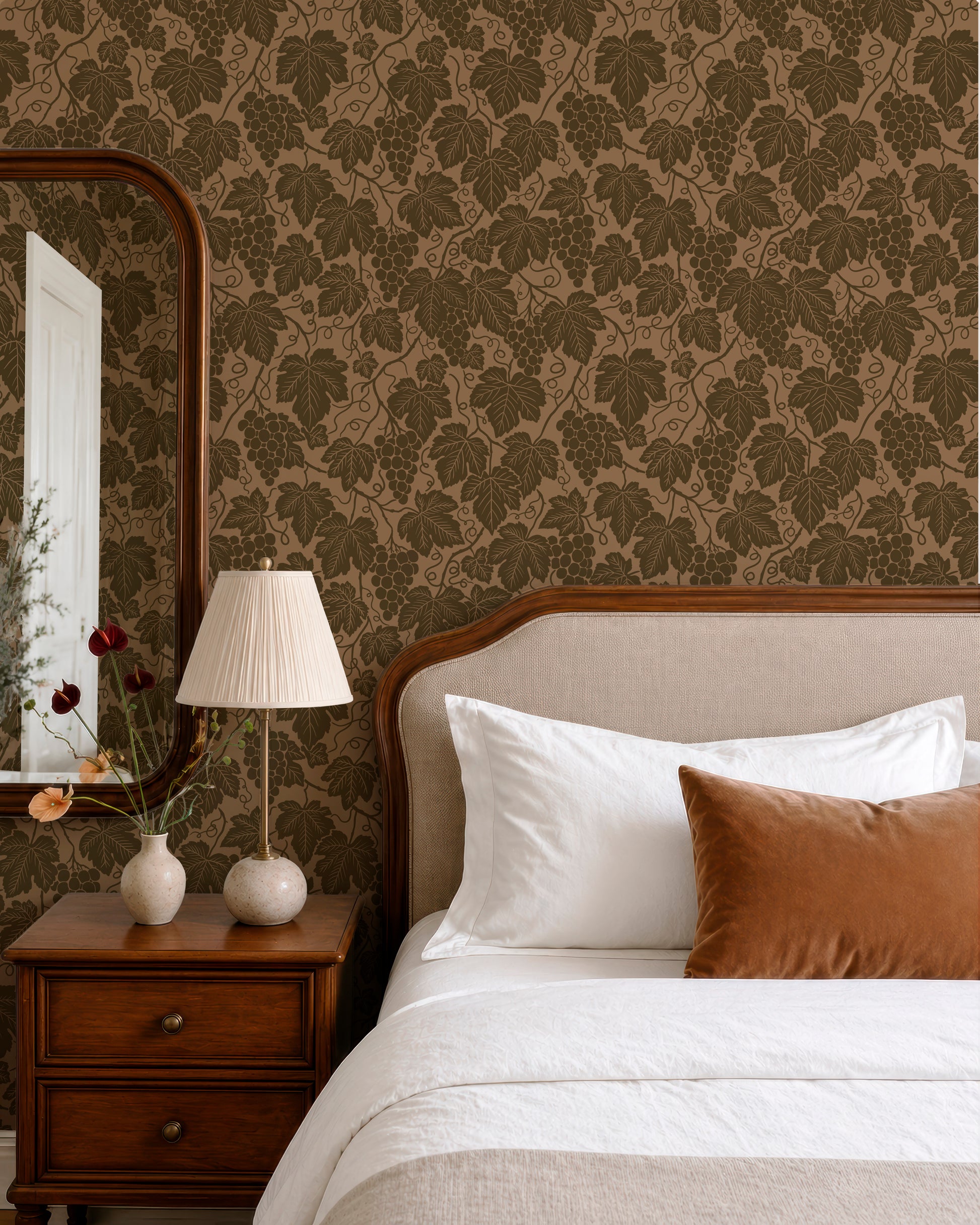 a bedroom with a bed, a nightstand, and a mirror. The wall behind the bed has a patterned wallpaper with leaves and grapes. The room has a warm and cozy atmosphere, with a brown and white color scheme.