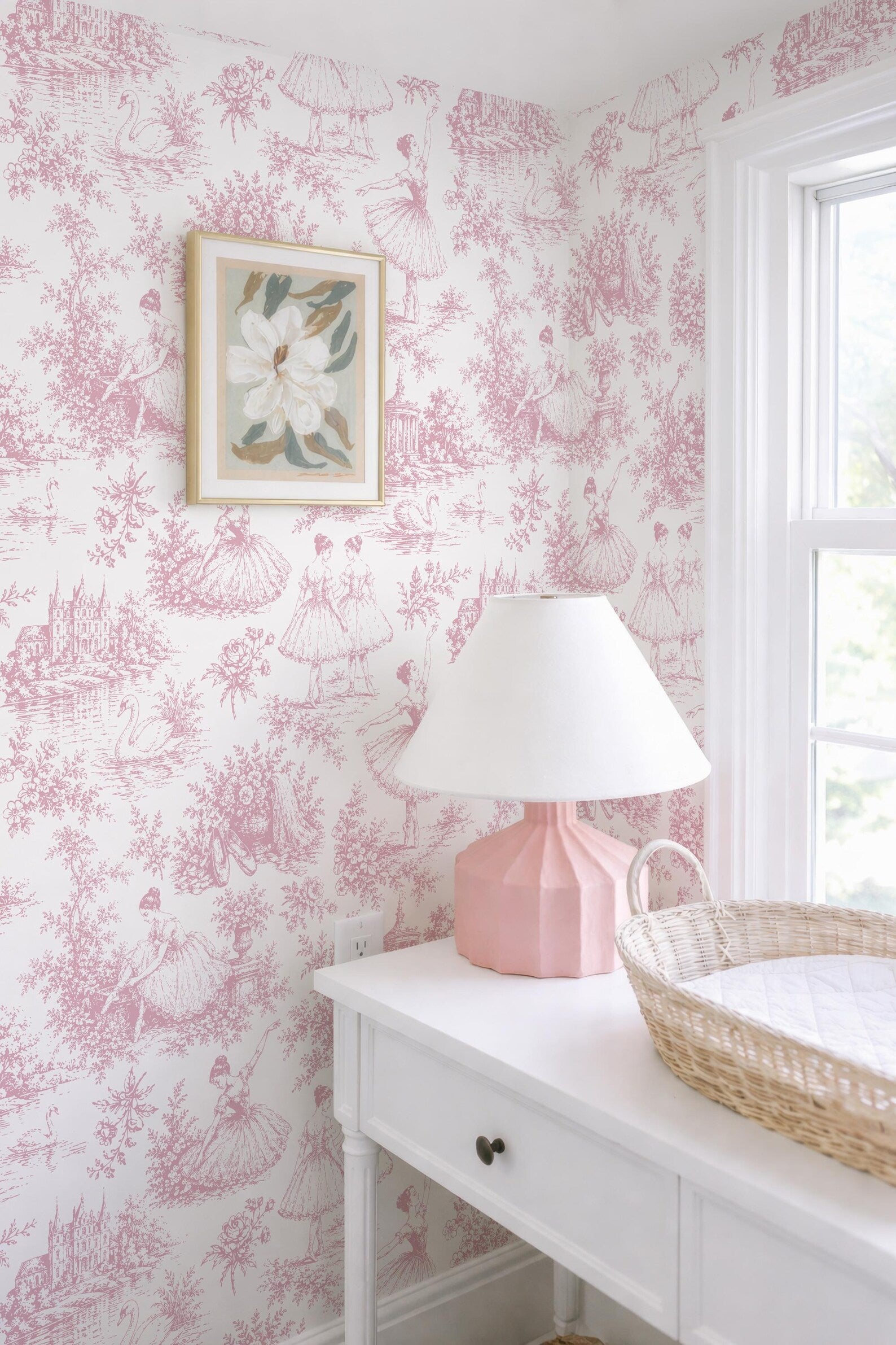 a pink and white color scheme with a white dresser, a pink lamp, and a woven basket. The dresser has a drawer and a mirror above it. The wall behind the dresser has a pink and white wallpaper with a floral pattern.