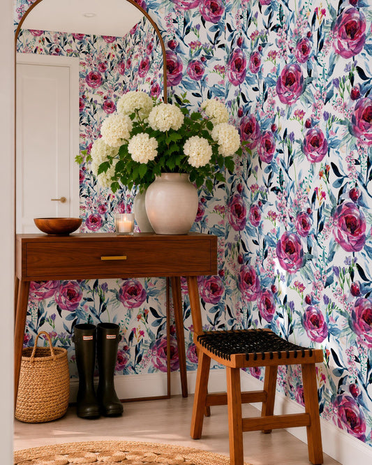 a room with a wooden table, a vase of white flowers, a mirror, a pair of black boots, and a woven basket. The walls are covered in a floral wallpaper with pink and white flowers.