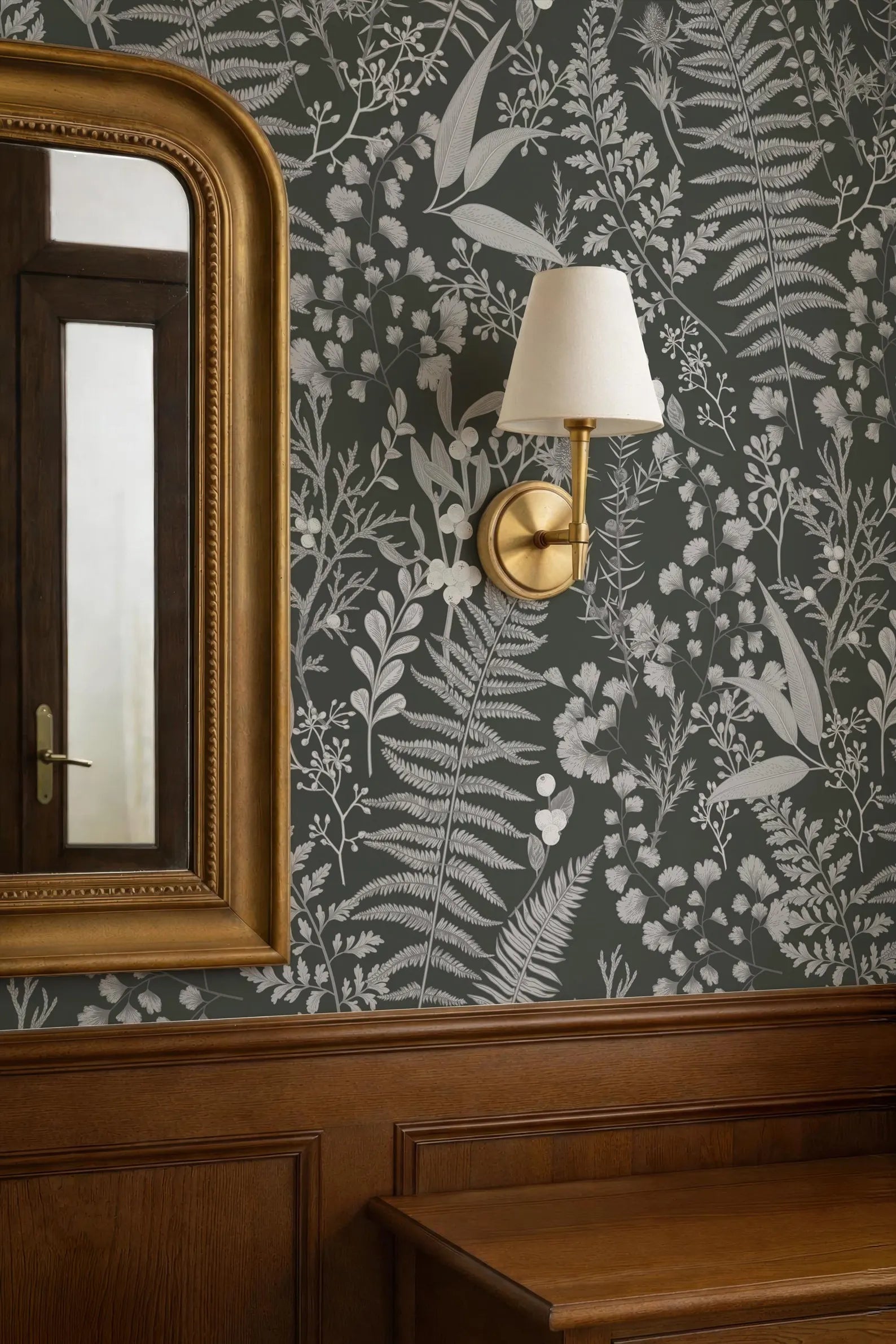 a wallpapered wall with a gold-framed mirror and a gold-framed wall lamp.