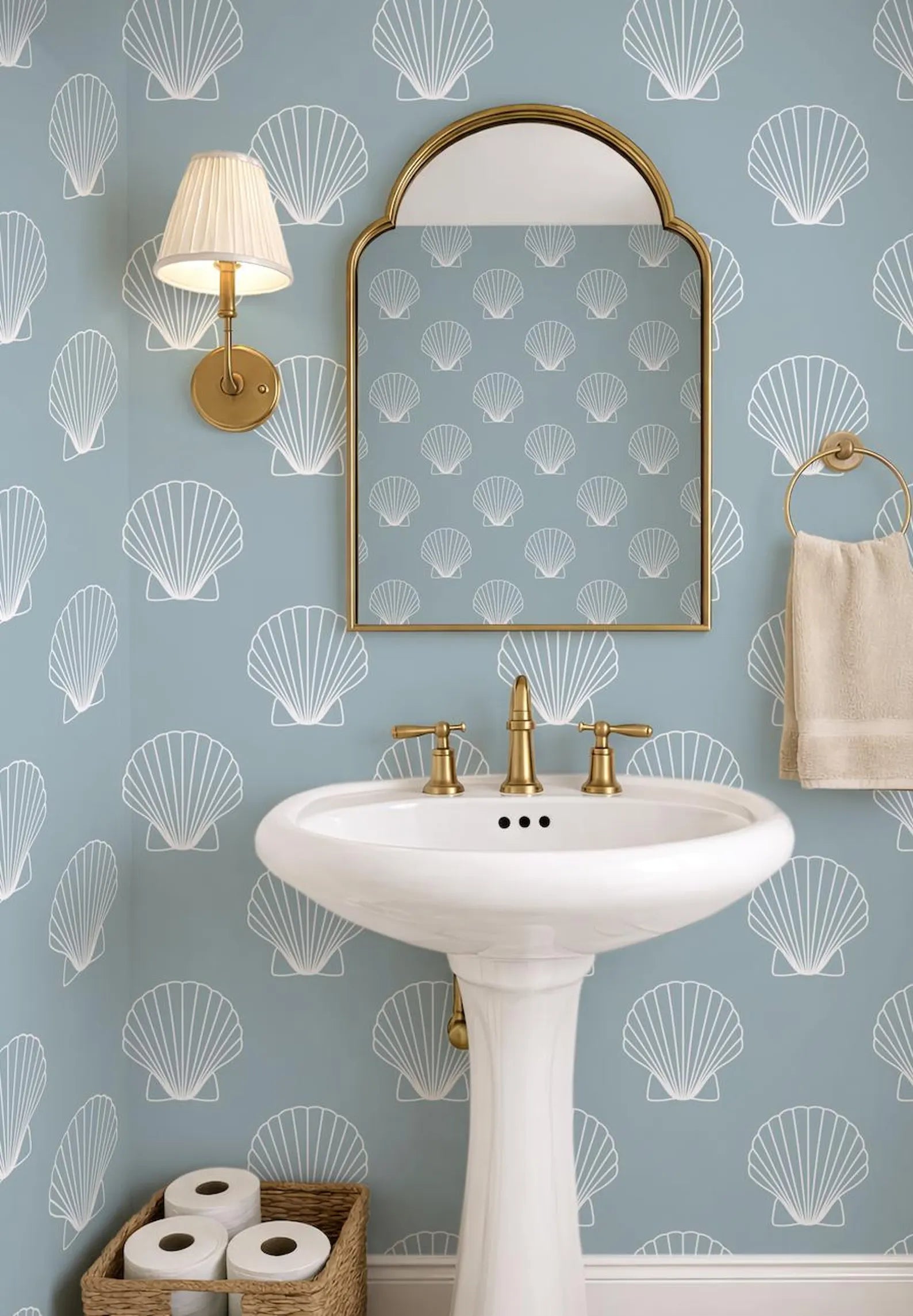 a bathroom with a white pedestal sink, a mirror with a gold frame, and a light fixture above the sink. The walls are covered in a pattern of white seashells, creating a coastal theme. The bathroom also includes a towel rack and a basket for toiletries.