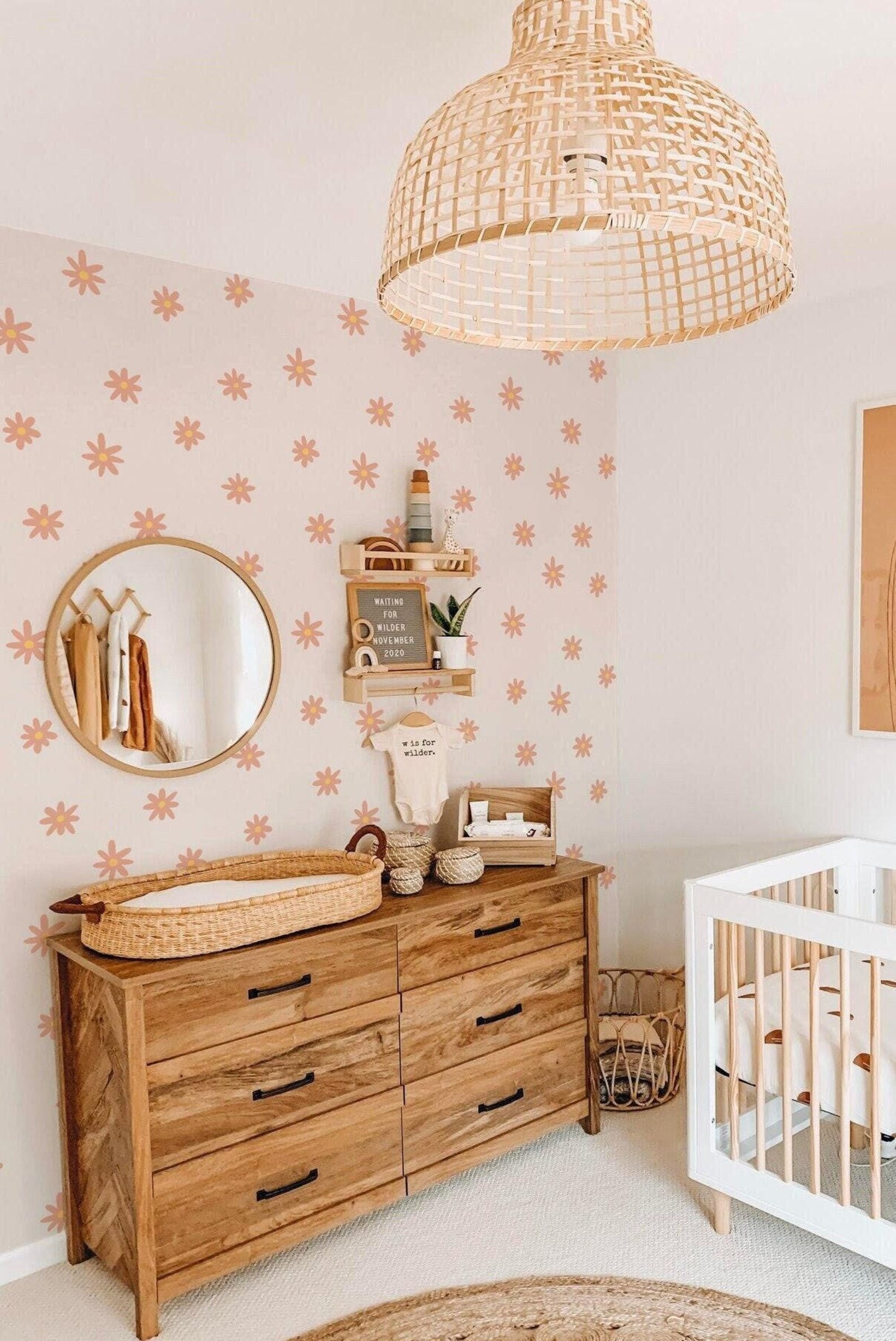 a cozy nursery with a wooden dresser, a round mirror, a woven light fixture, and a white crib. The walls are decorated with orange and white floral patterns, creating a warm and inviting atmosphere.